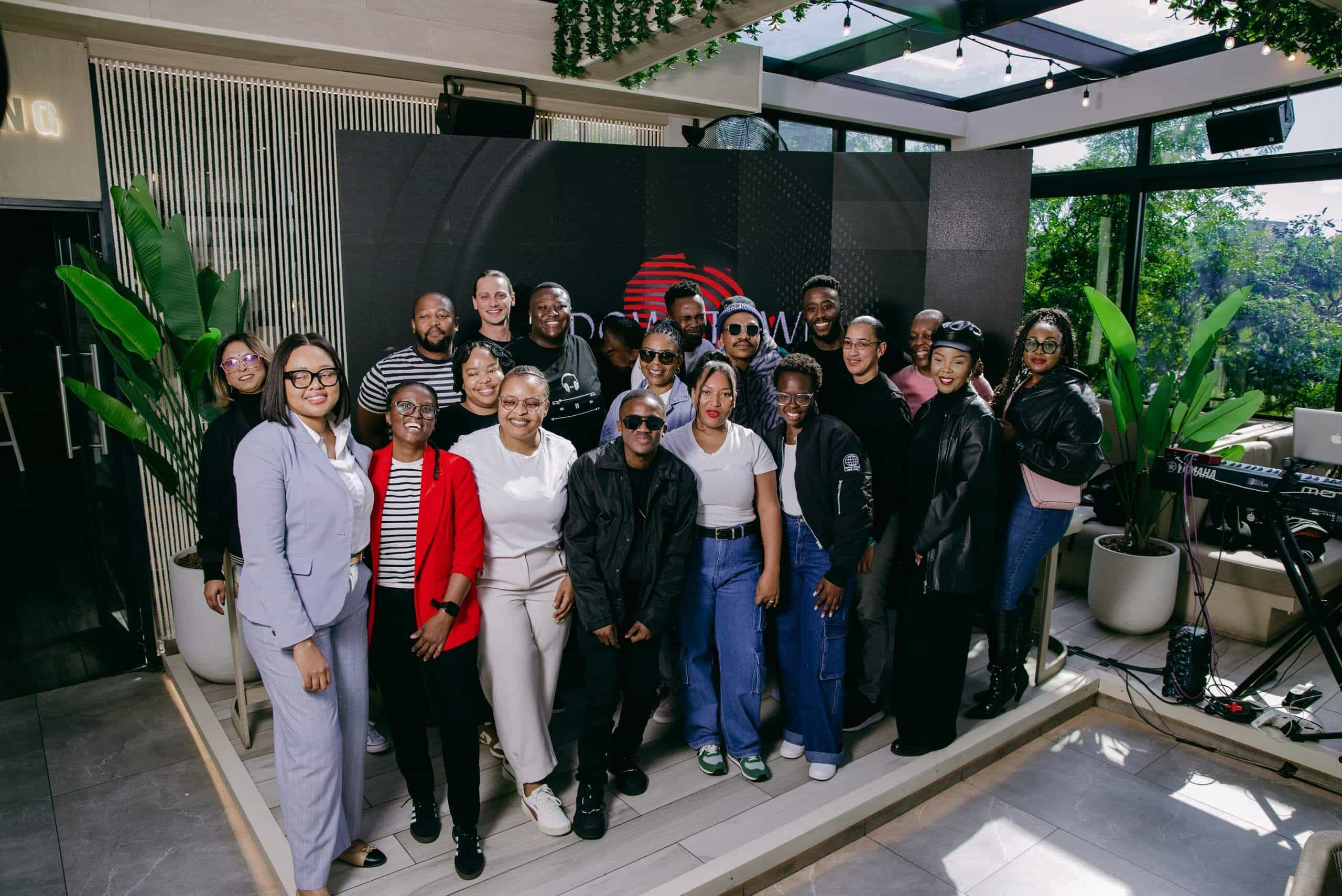 A diverse group of about 20 people, dressed in casual and business attire, poses and smiles together in a modern, plant-filled indoor space with large windows and greenery. A digital display and musical equipment appear in the background.