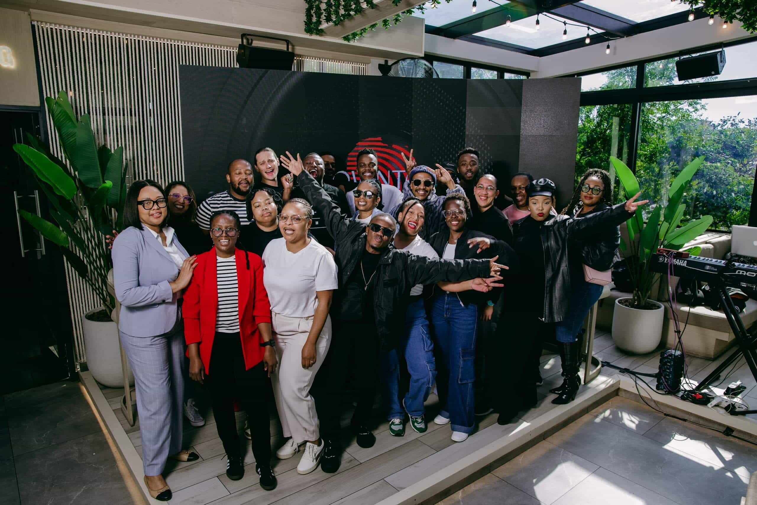 A diverse group of people pose cheerfully together indoors, with some raising their arms or making peace signs. They stand on a small stage with plants and a keyboard nearby, in a sunlit room with large windows and modern decor.