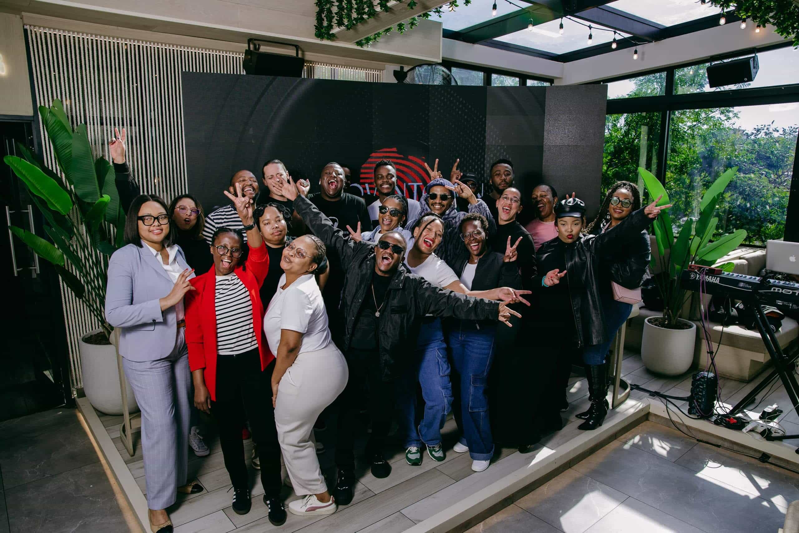 A large group of people, diverse in age and style, pose together indoors, many making peace signs or playful gestures. Some wear sunglasses and dark outfits. There are plants and a keyboard in the background, with daylight streaming through large windows.