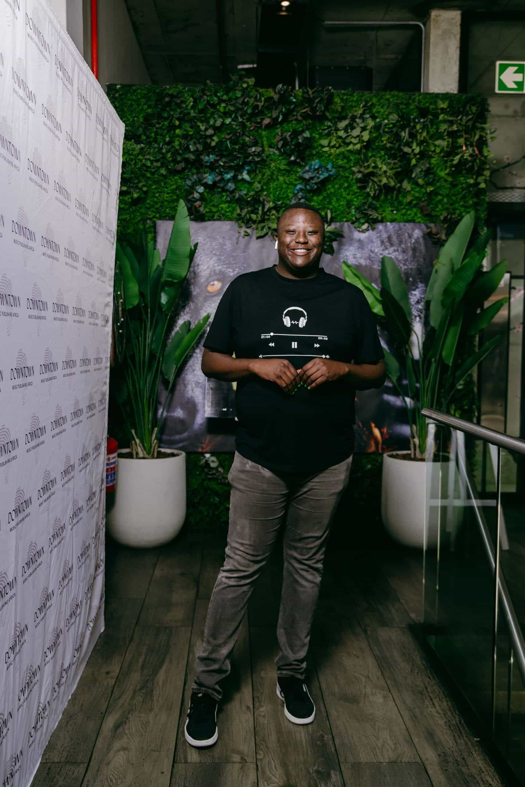 A smiling man stands indoors on wooden flooring, wearing a black T-shirt with white graphics, gray pants, and black sneakers. Behind him are green plants in white pots and a lush vertical garden wall, with a branded white backdrop to the left.