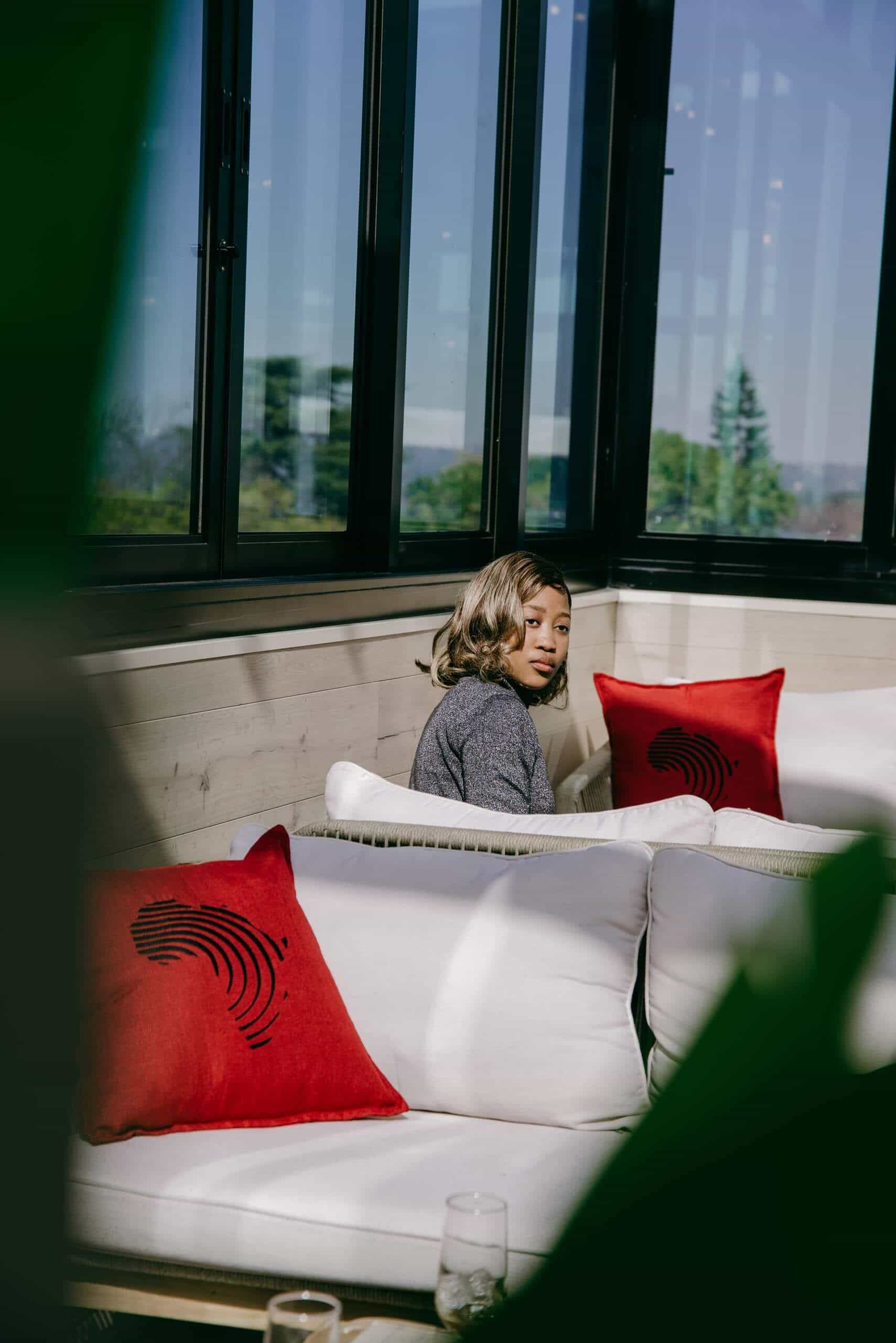 A woman with shoulder-length hair sits on a white couch in a sunlit room with large glass windows. Red pillows with black designs are on the couch. Green leaves are blurred in the foreground, and trees are visible through the windows.