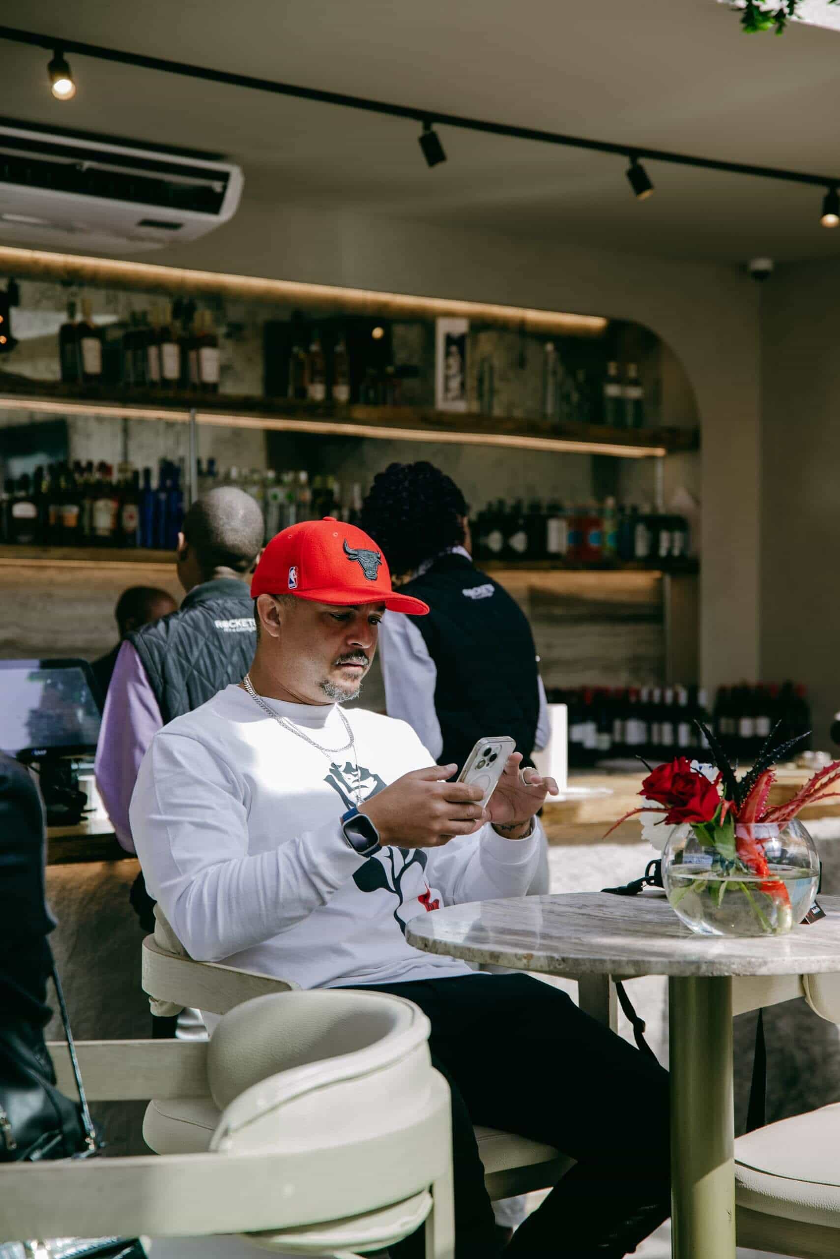 A man wearing a red Chicago Bulls cap and white sweatshirt sits at a marble table in a bar, looking at his phone. Behind him are shelves of bottles and other people. On the table is a glass vase with red and black flowers. The setting appears casual and modern.