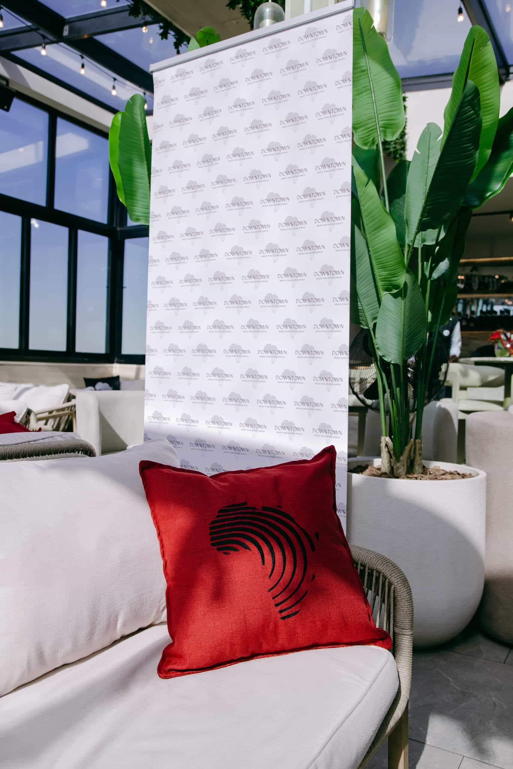 A red pillow with a black abstract pattern sits on a light-colored sofa. Behind it is a banner with repetitive gray logos, and large potted plants. The setting appears to be a modern, sunlit indoor lounge with big windows.