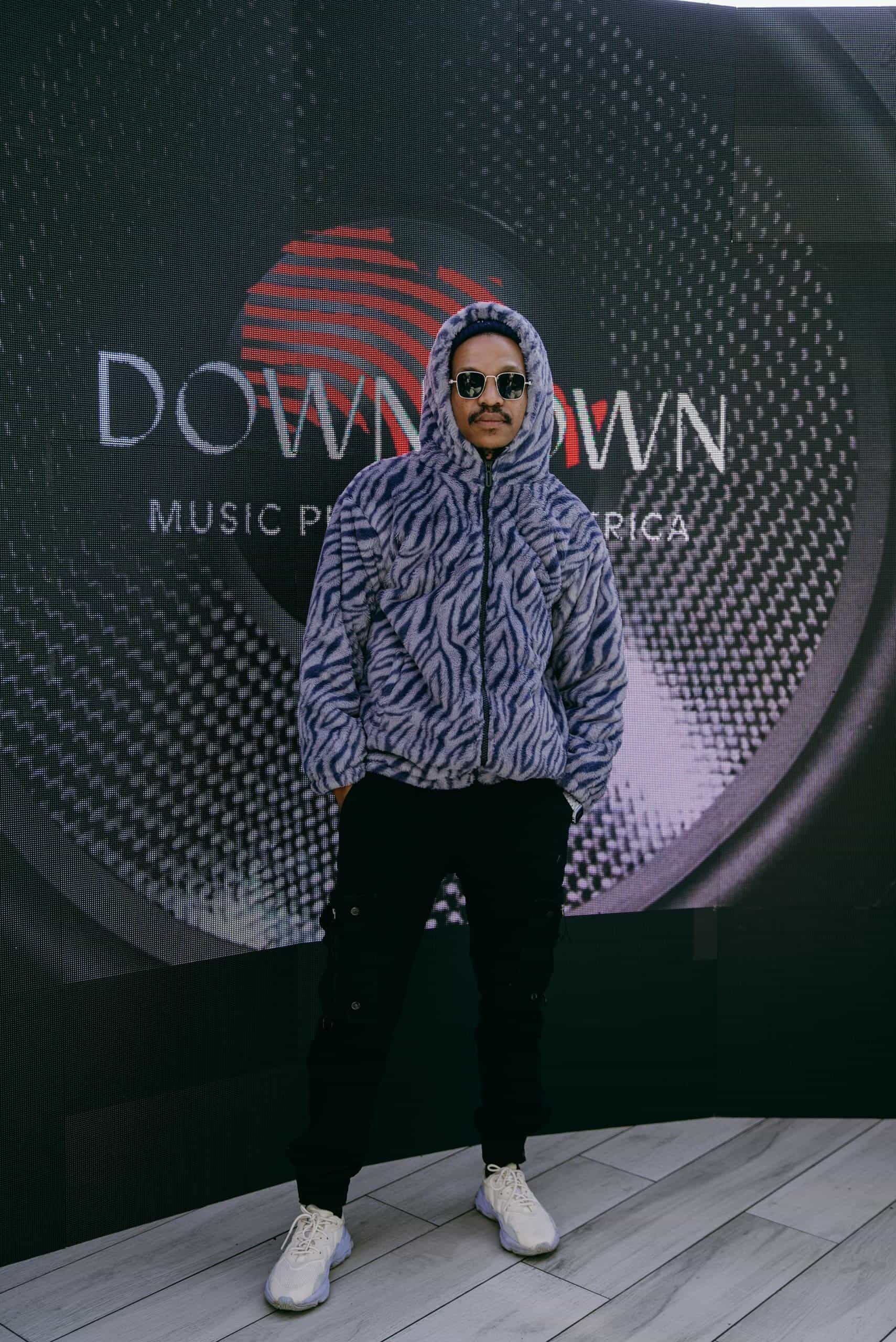 A man wearing a gray and black zebra-print hoodie, black pants, white sneakers, and sunglasses stands with hands in pockets in front of a large sign that reads 