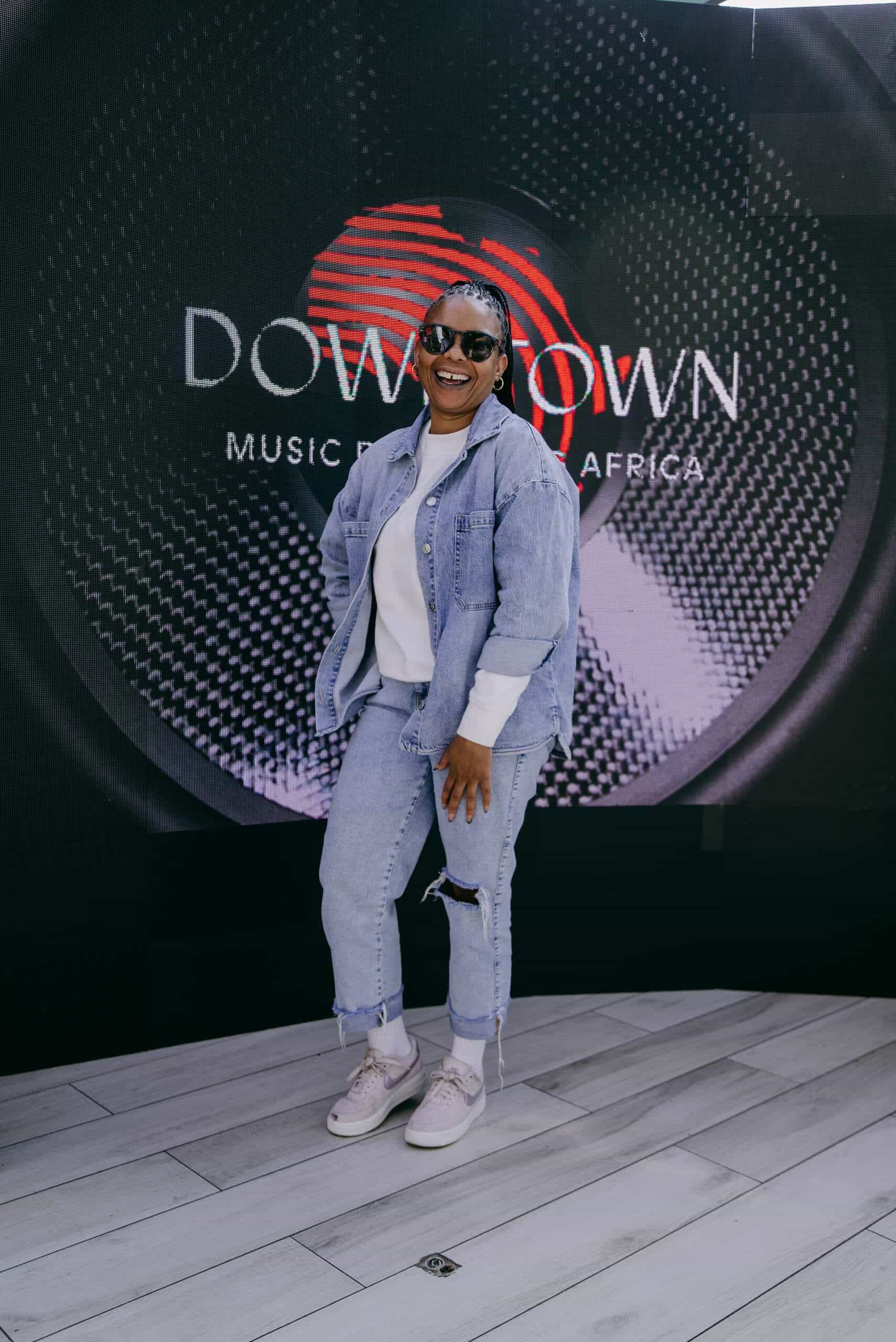 A smiling person wearing sunglasses, a white shirt, denim jacket, and ripped jeans poses confidently in front of a large 