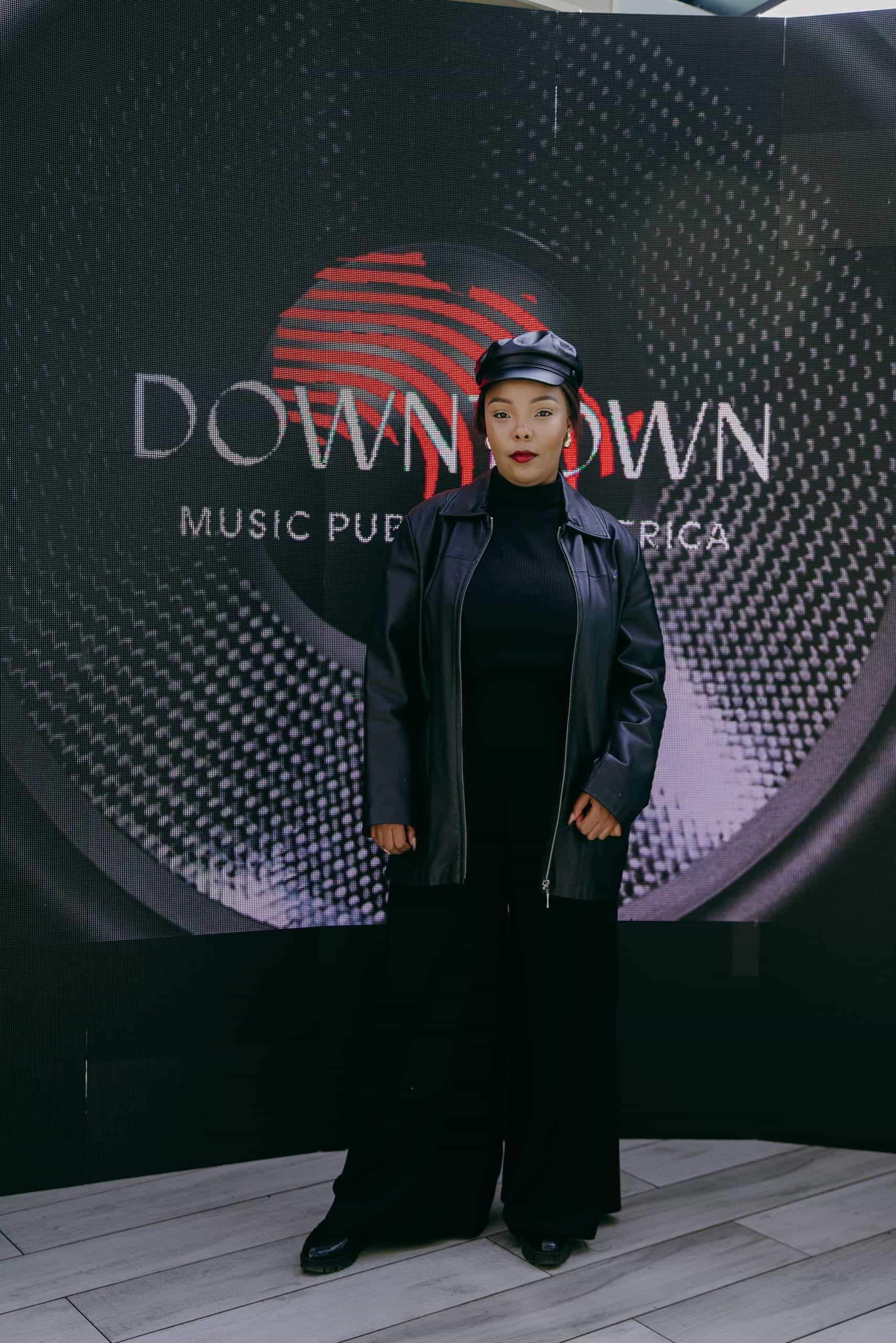 A woman in a black leather beret, black turtleneck, and black leather jacket stands confidently in front of a large circular graphic backdrop that reads &ldquo;DOWNTOWN Music Publishing Africa&rdquo; with abstract patterns resembling a vinyl record.