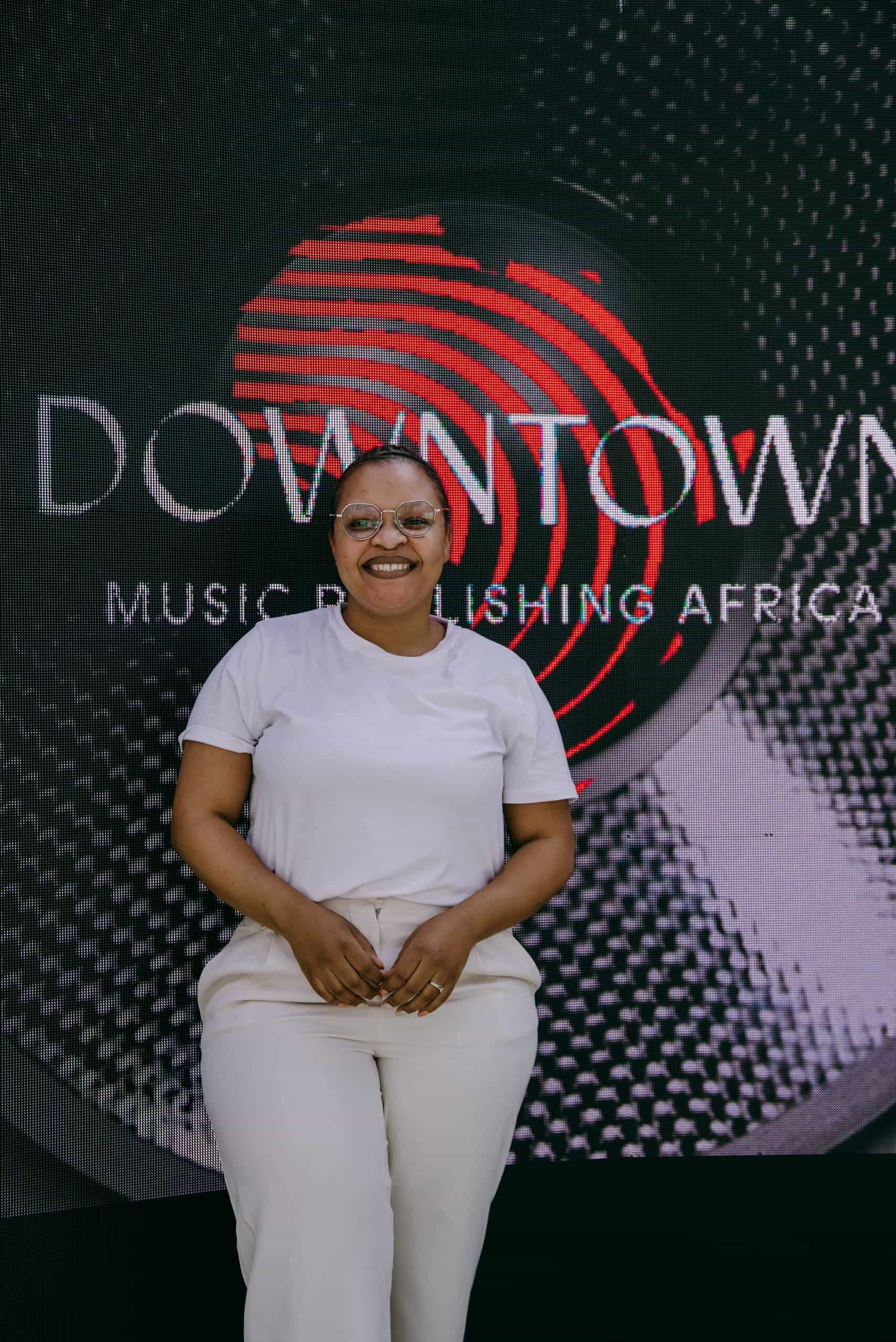 A smiling person in glasses and a white outfit stands in front of a large black backdrop with a red fingerprint and the words &ldquo;DOWNTOWN MUSIC PUBLISHING AFRICA&rdquo; printed on it. The person appears relaxed and confident, with hands clasped in front.