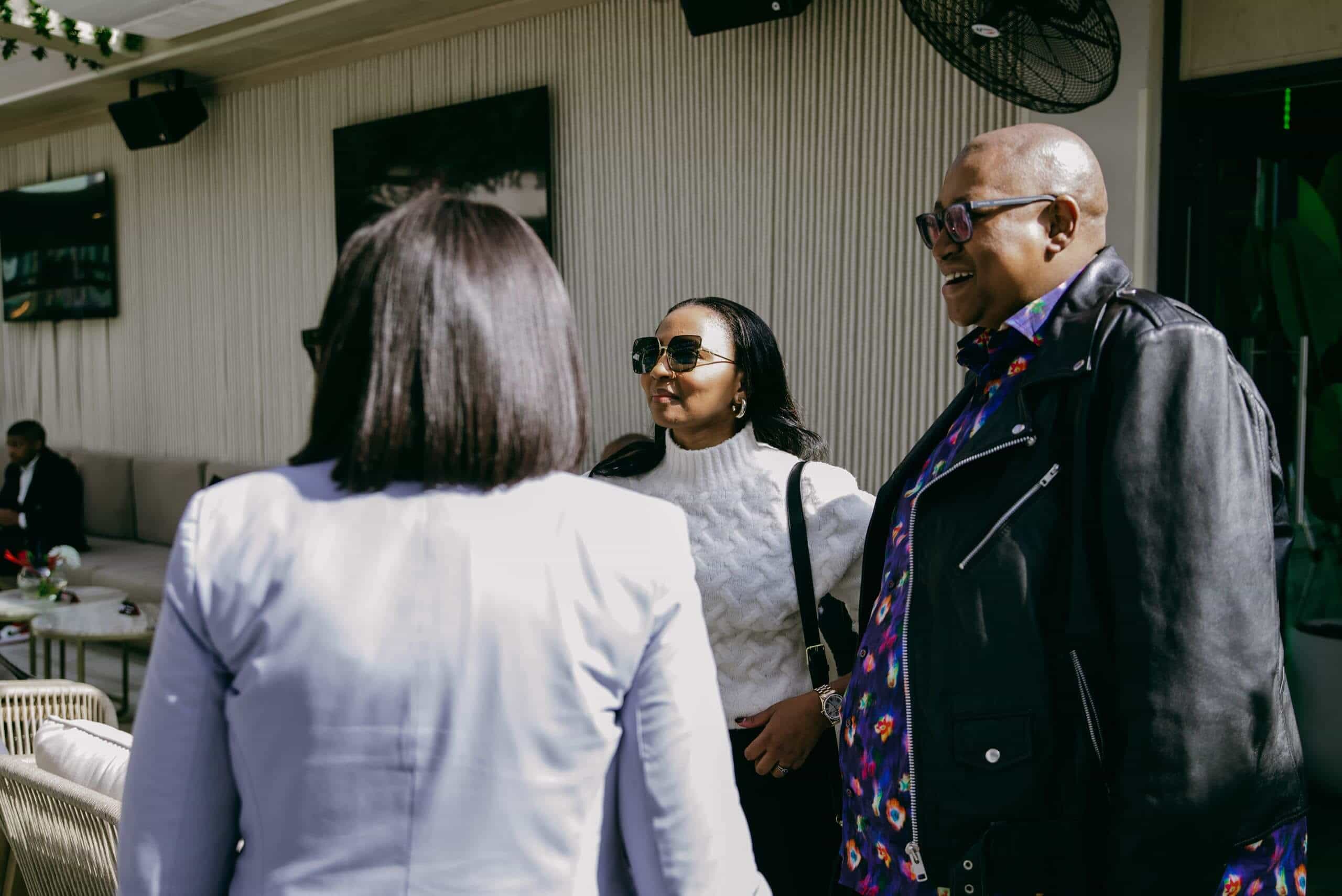 Three people stand outdoors, chatting and smiling. One, in a bright patterned shirt and black jacket, laughs; another wears a white sweater and sunglasses. The third, seen from behind, has straight shoulder-length hair and a light blazer. Patio seating and decor are visible in the background.