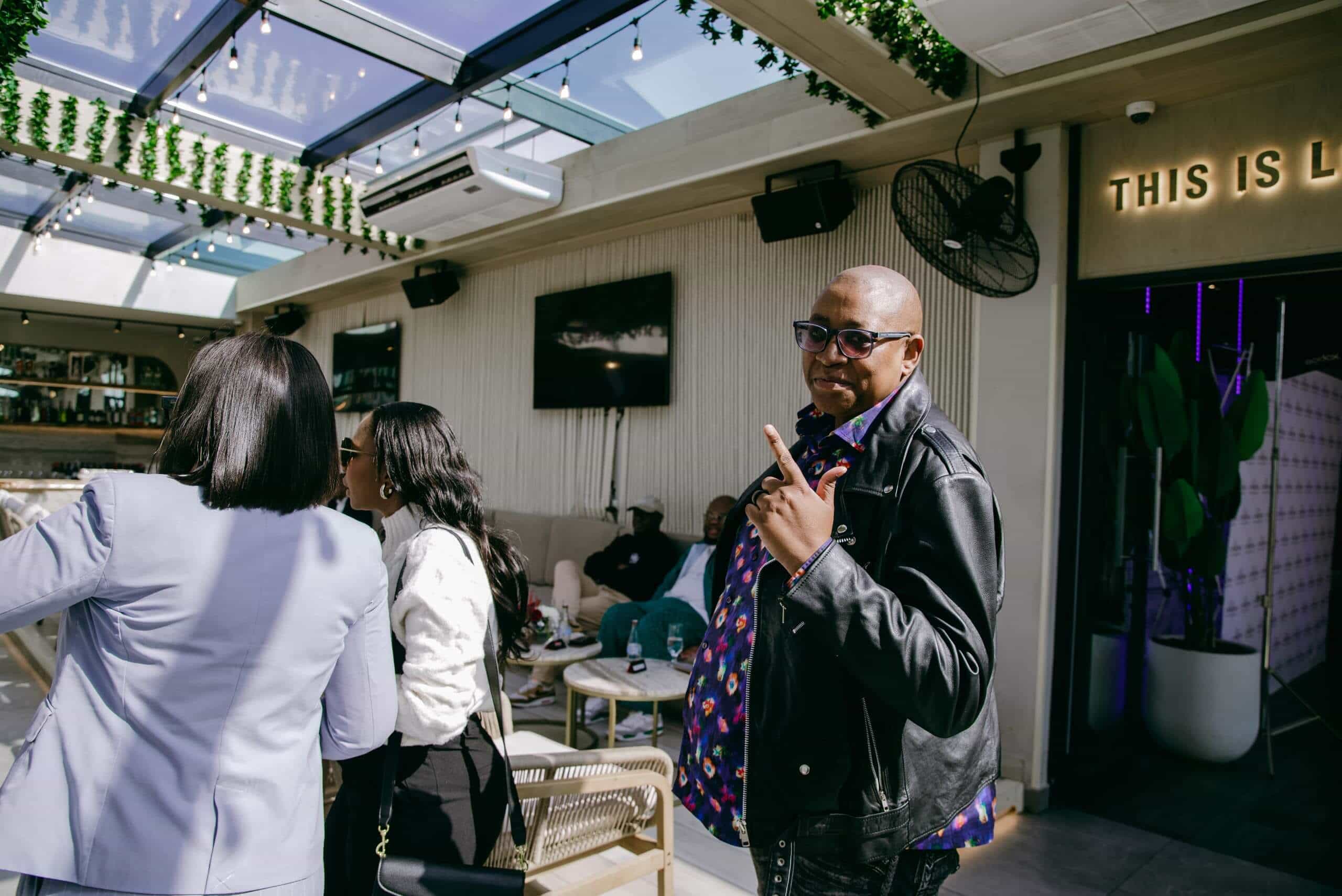 A man in sunglasses and a black leather jacket makes a peace sign at the camera in a stylish indoor lounge with a glass ceiling. Two women with their backs to the camera talk nearby. People sit and relax on sofas in the background.