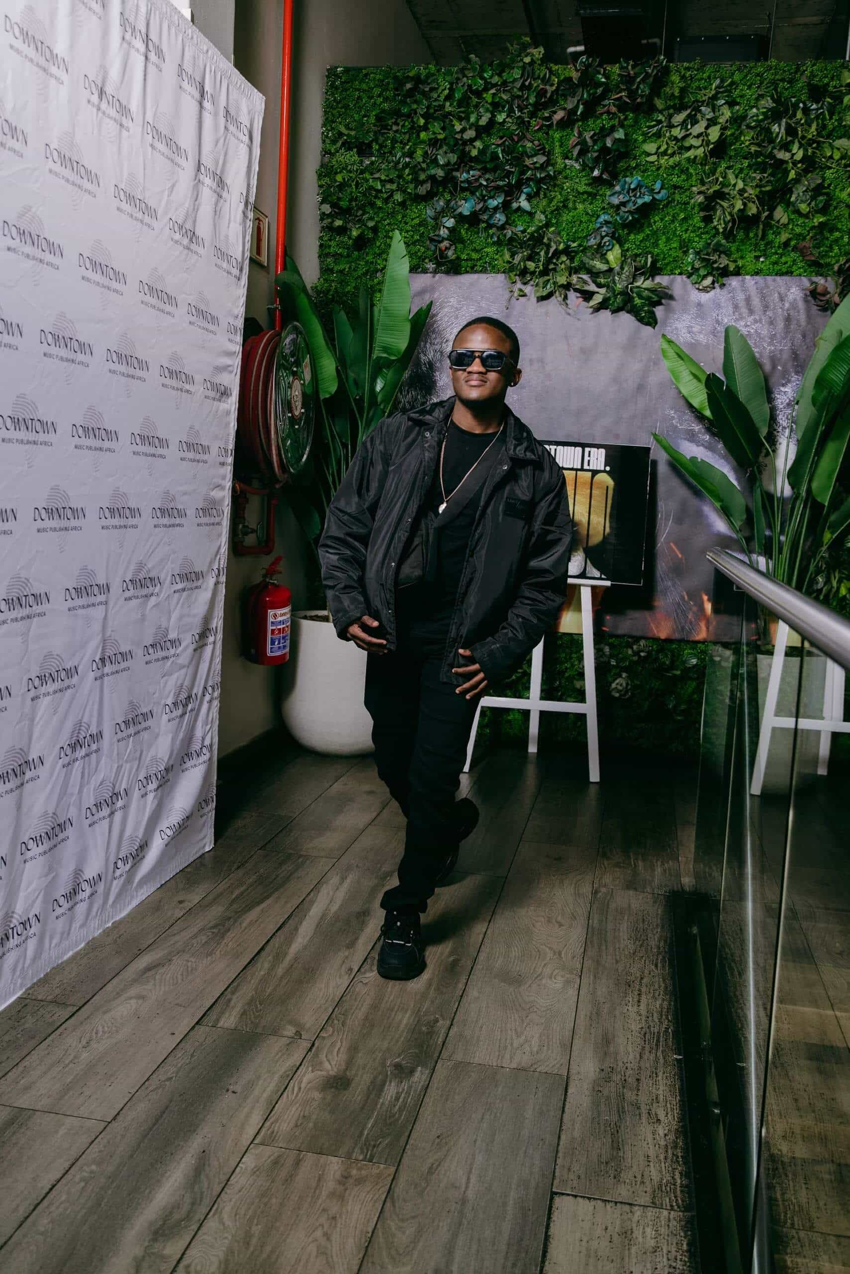 A person in black sunglasses, jacket, pants, and sneakers poses indoors. They lean against a glass railing, one leg crossed over the other. There are green leafy plants, a fire hose, poster, and a wall covered in greenery in the stylish, modern setting.