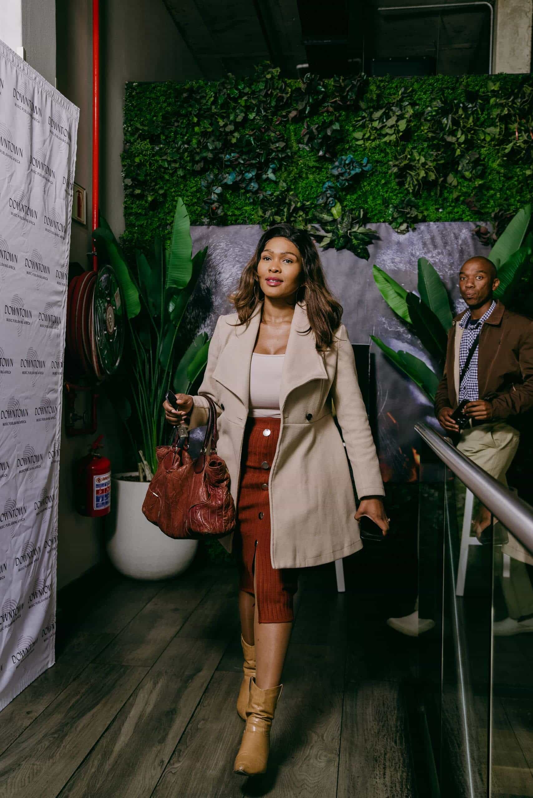 A woman in a beige coat, rust skirt, and tan boots walks confidently indoors, holding a brown handbag. She has shoulder-length hair and a calm expression. Behind her, a man in a jacket stands by a green plant wall, near a red fire extinguisher.