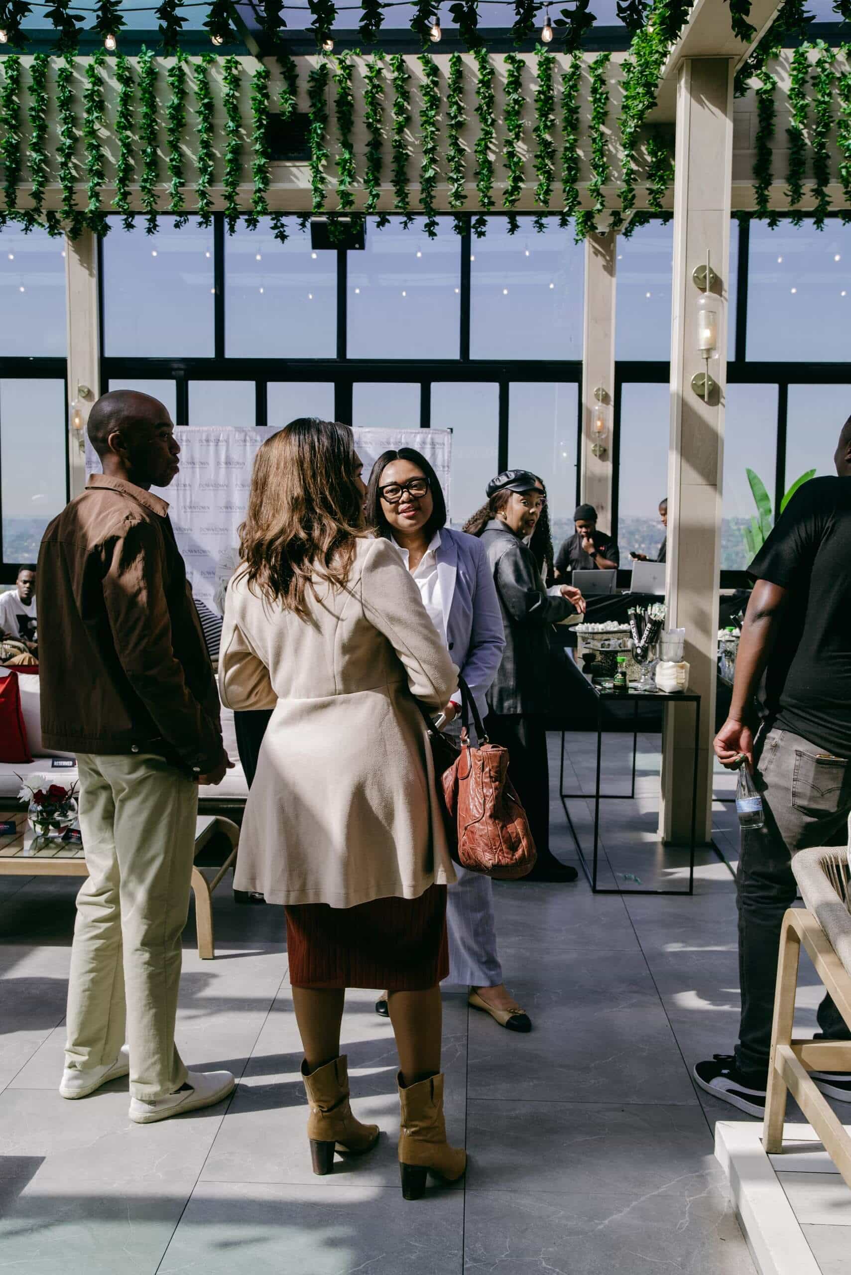 A group of people chat indoors at a sunlit, modern venue with tall windows, leafy decor above, and string lights. A woman in a beige coat and boots faces others, including a woman in a light suit and glasses, and a man in a dark jacket.