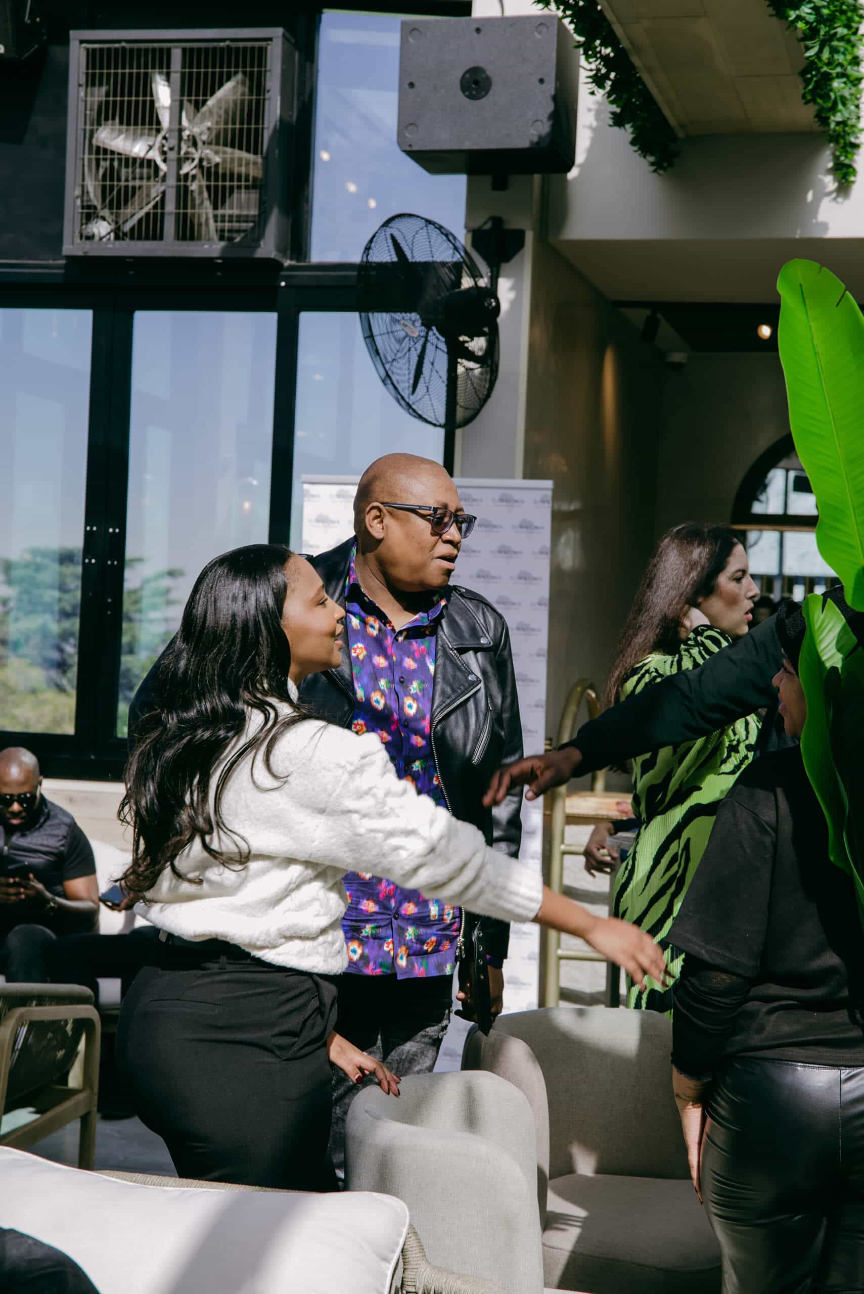 Three people greet each other in a bright indoor space with large windows and tall plants. A man in glasses and a black leather jacket stands between two women, one in a white sweater and another in black, as they reach out for a hug or handshake.
