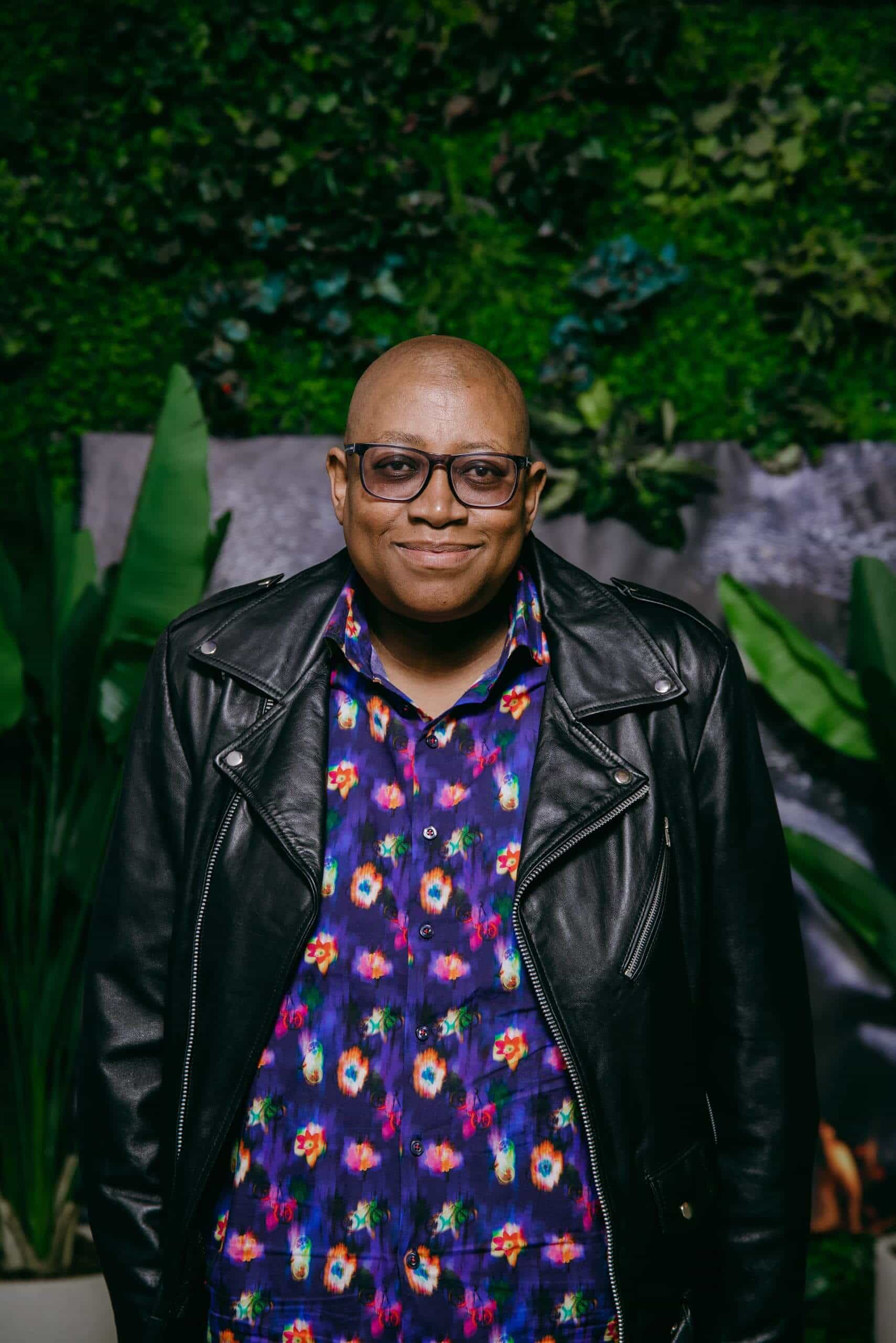 A smiling person with glasses and a shaved head wears a black leather jacket over a colorful, floral-patterned shirt. They stand in front of a lush green backdrop with various plants and leaves, creating a vibrant, natural setting.