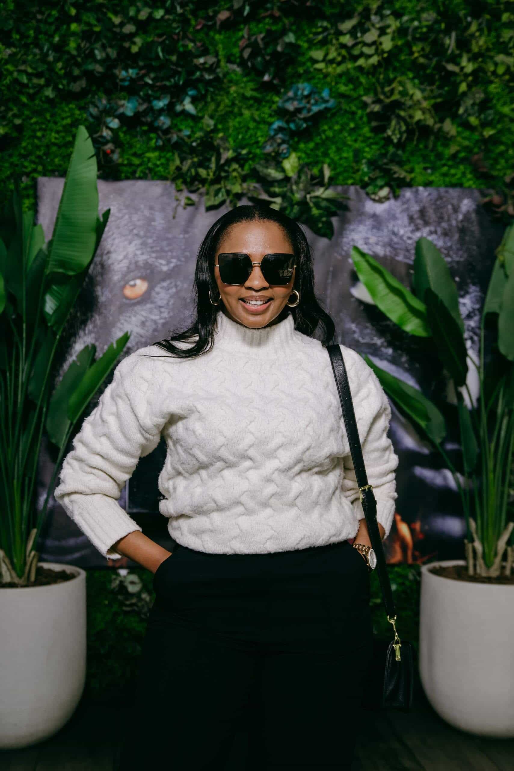 A woman with long dark hair wears sunglasses, a white cable-knit turtleneck sweater, and black pants. She stands with hands in pockets, smiling, in front of a green leafy backdrop with large potted plants and a blurred mural behind her.