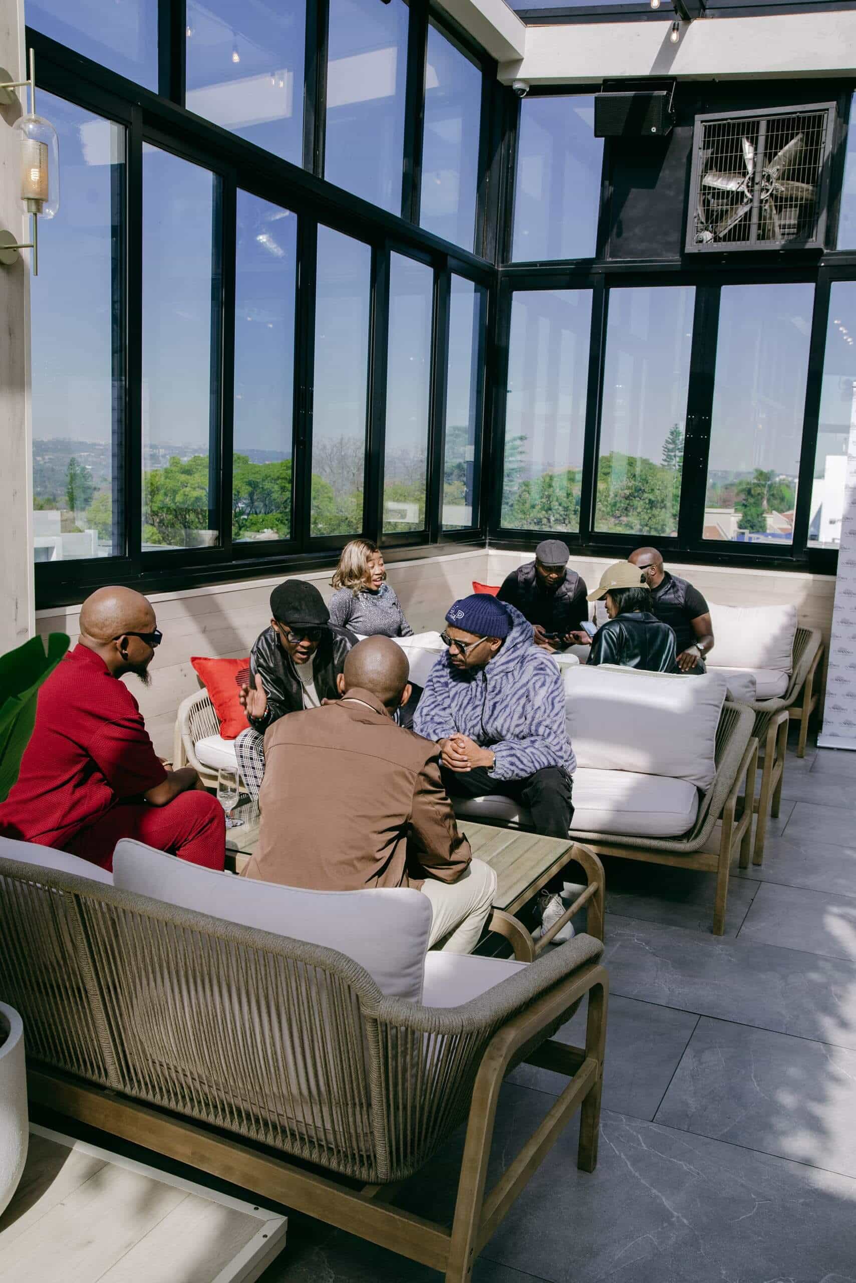 A group of older adults sit in conversation on cushioned chairs in a bright, glass-enclosed lounge with large windows. Sunlight fills the modern space, and greenery is visible outside. The mood appears relaxed and social.