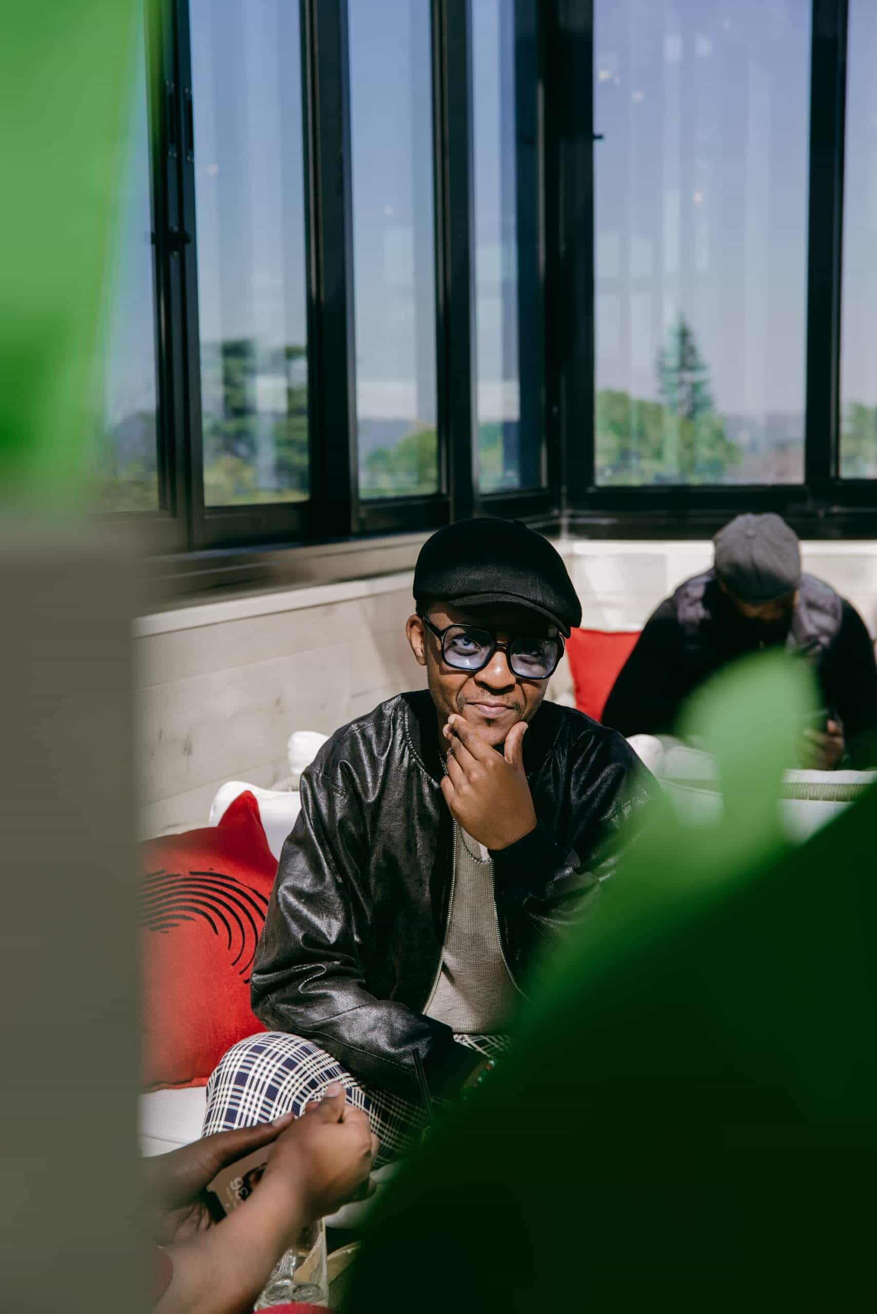 A man wearing glasses, a black cap, and a leather jacket sits on a white couch with red pillows, touching his chin and smiling. Another person is seated in the background, partially obscured, and green leaves frame the foreground.