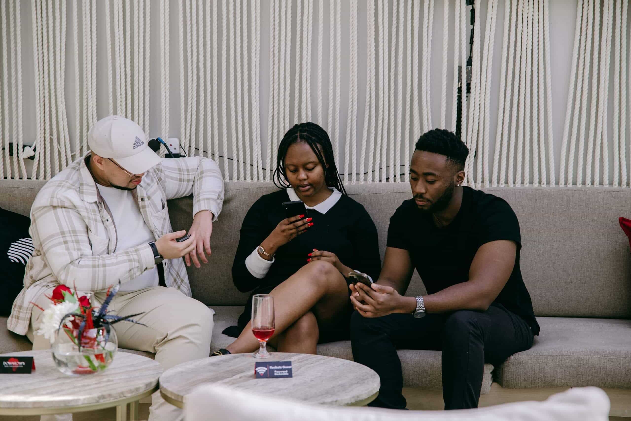 Three people sit on a beige couch, focused on their phones. The person on the left wears a white cap and plaid jacket, the middle person wears a black dress, and the right person wears a black t-shirt. A red drink and flowers are on the table in front of them.