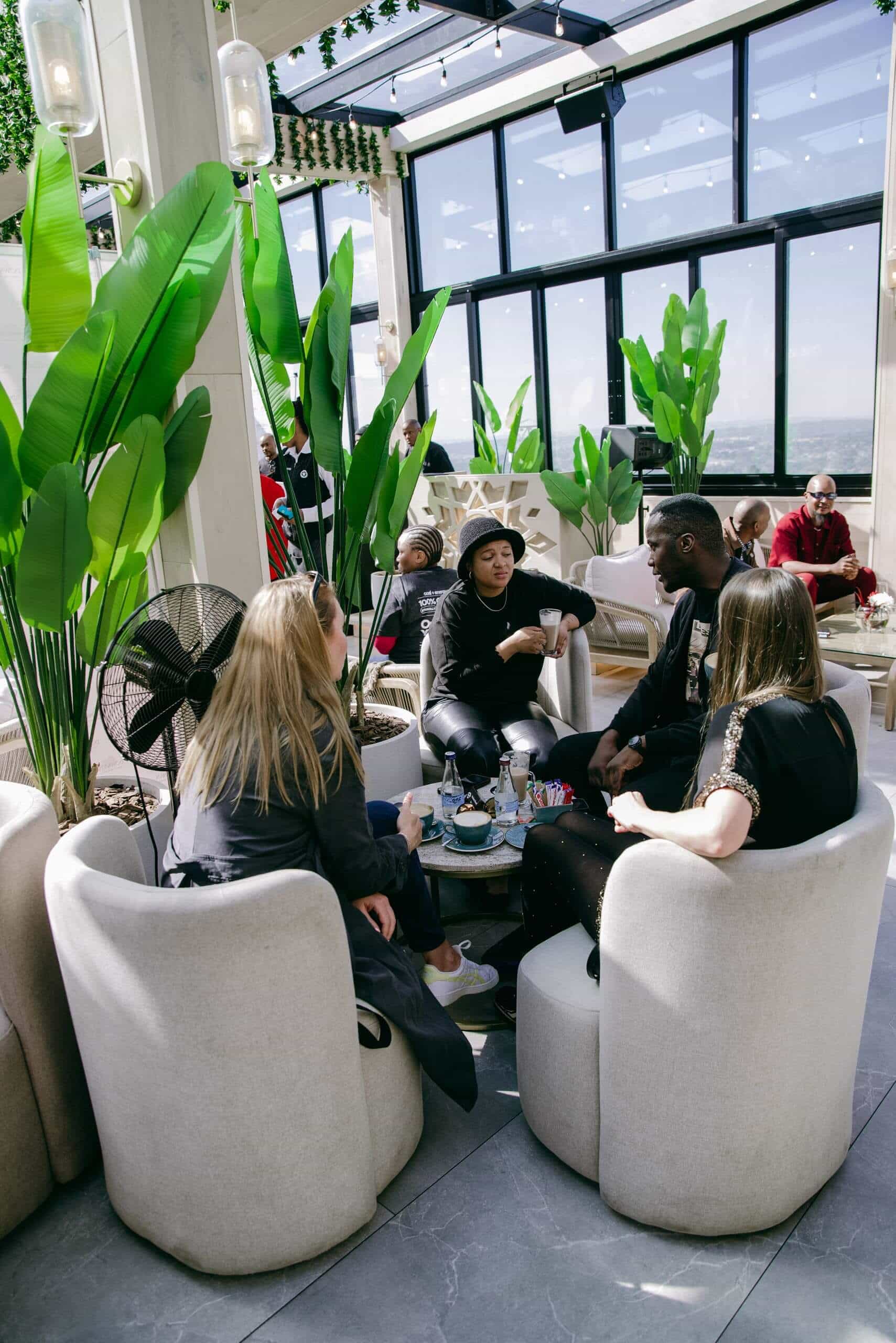 A group of five people sit in a modern, sunlit lounge with large windows and lush green plants. They are engaged in conversation, seated in a circle of white cushioned chairs around a small coffee table with drinks. Other people are visible in the background.