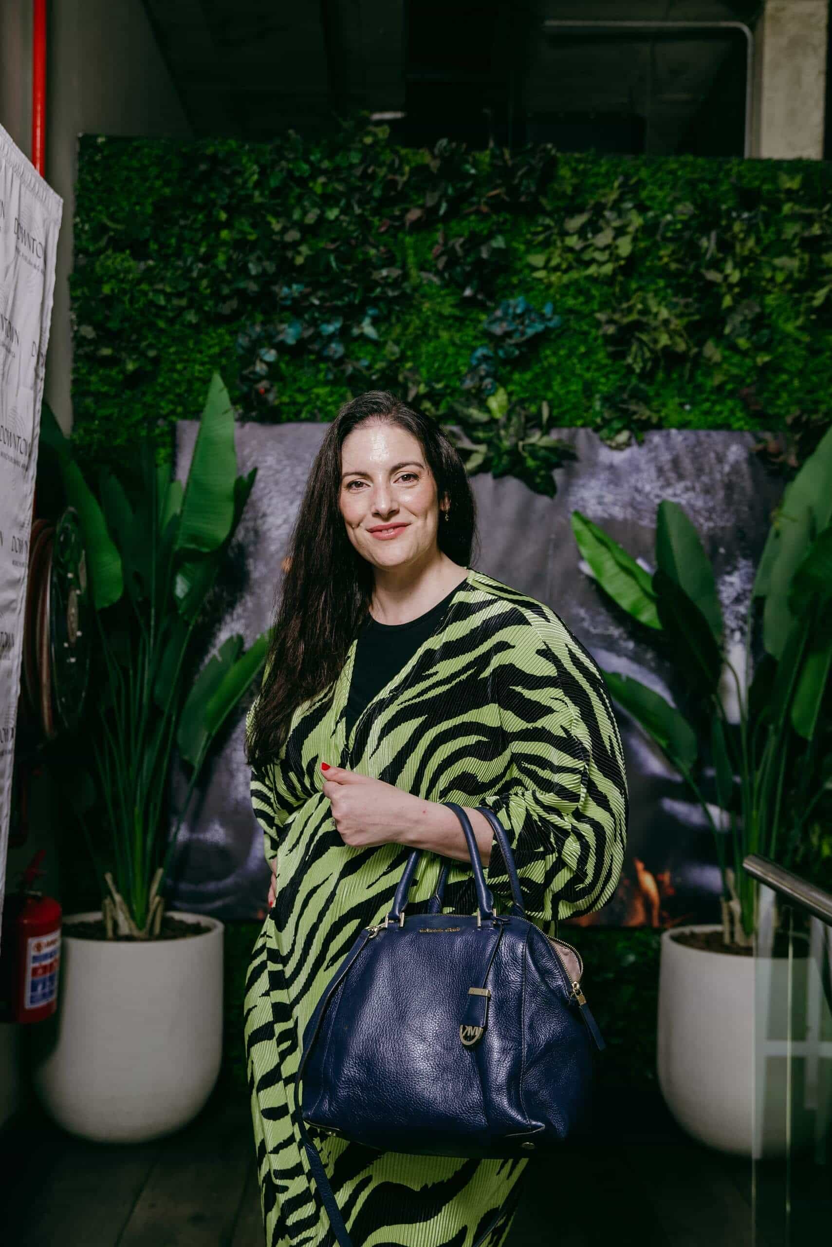 A woman with long dark hair, wearing a green and black patterned dress, stands indoors in front of a lush green leafy wall. She holds a large navy blue handbag and smiles slightly. Two potted plants are positioned on either side of her.