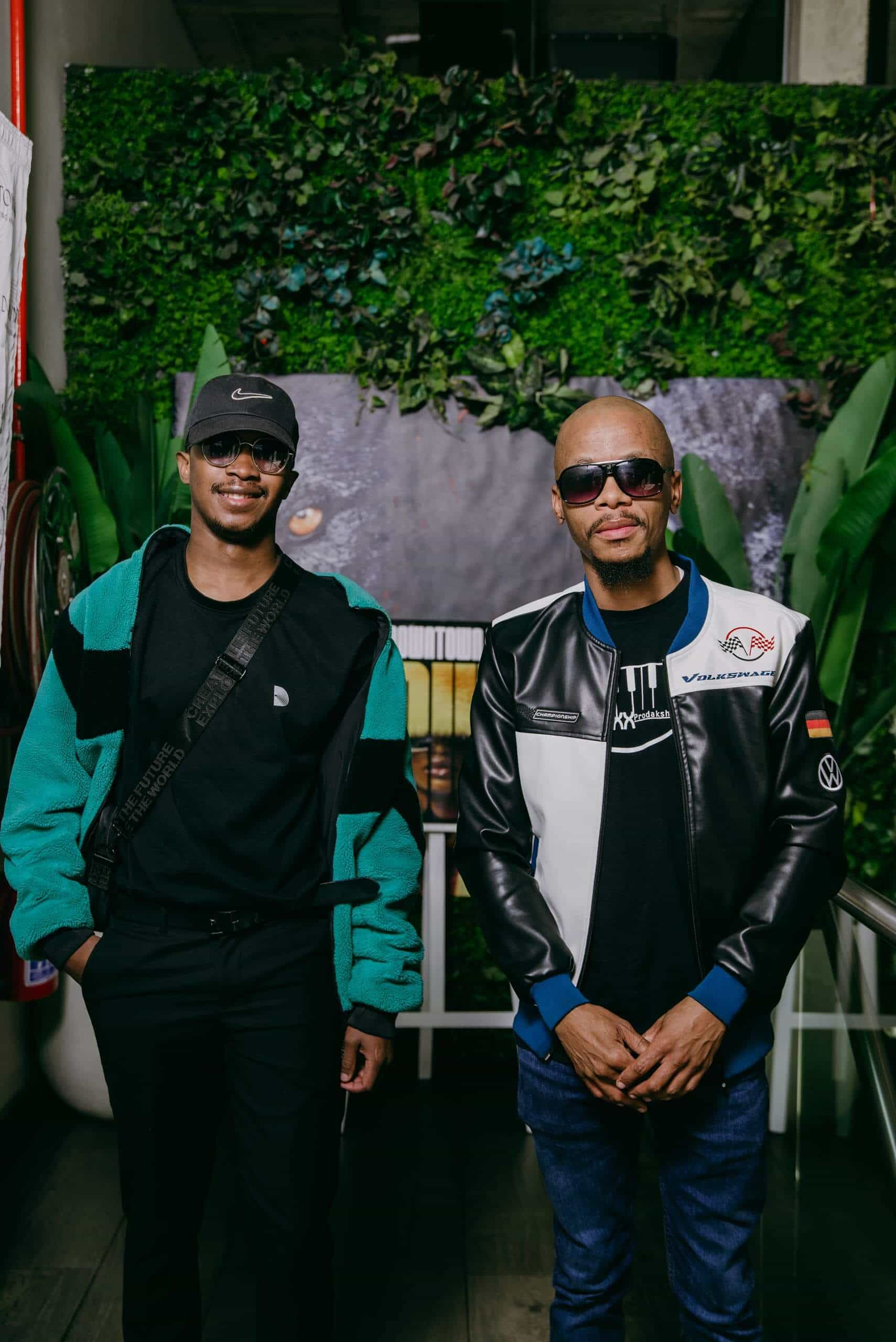 Two men stand side by side indoors in front of a leafy green wall and animal mural. Both wear sunglasses. The man on the left has a teal and brown jacket, black pants, and a cap. The man on the right wears a black-and-white leather jacket and blue shirt.