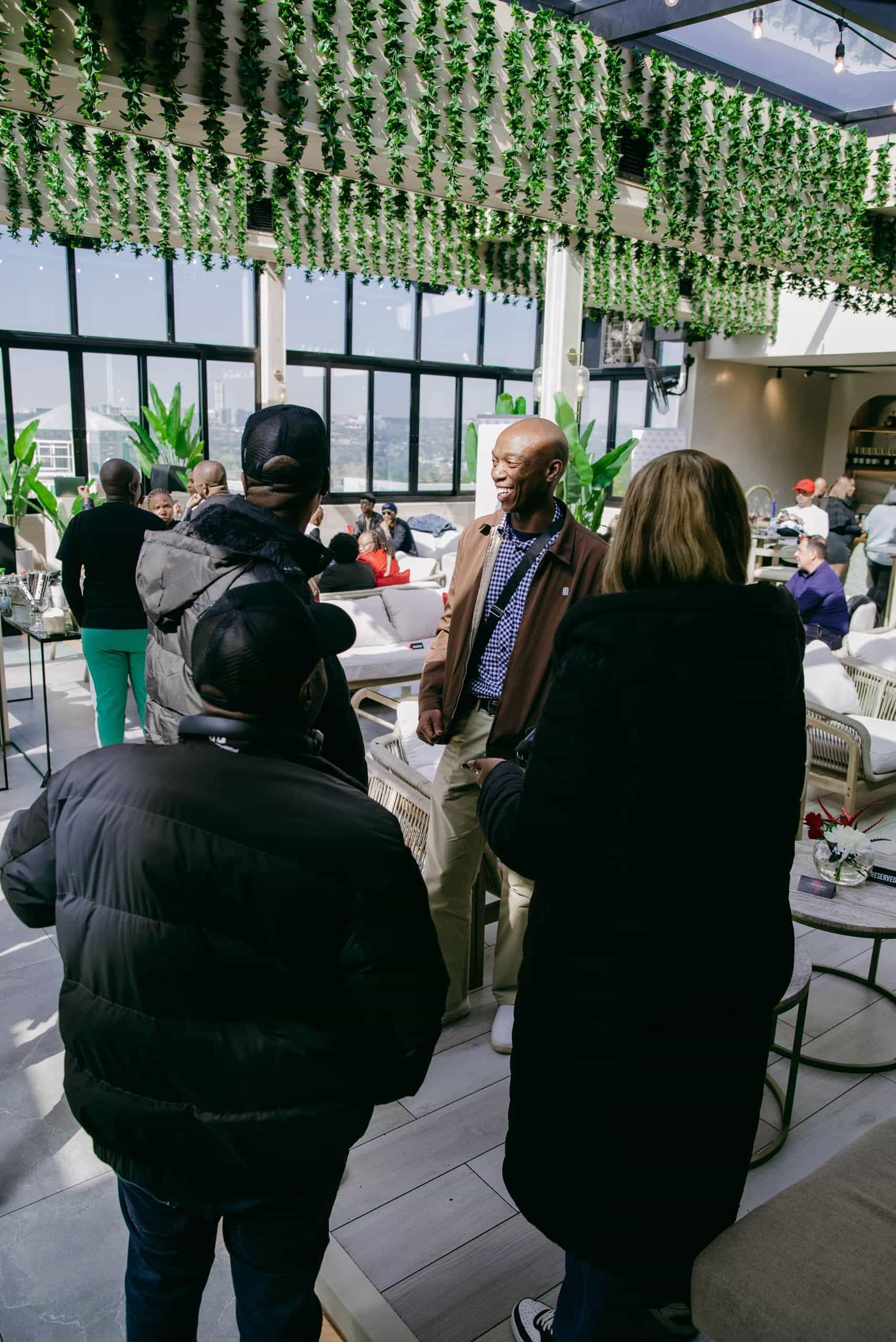 A group of people, mostly in dark jackets, stand indoors talking in a modern, sunlit lounge. One man in a brown jacket and scarf faces the group, smiling. The space features large windows, green hanging plants, and people seated in the background.