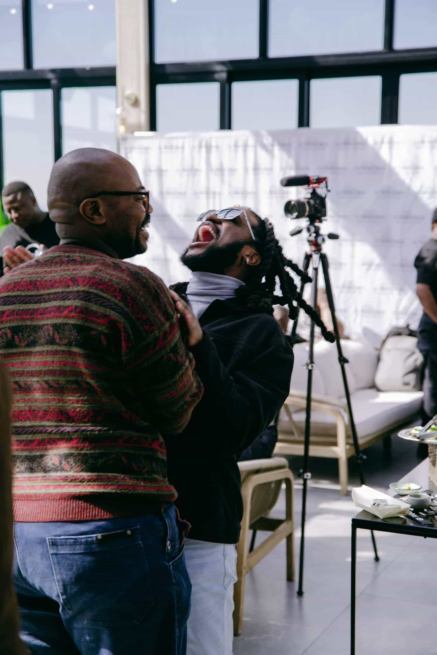 Two people stand laughing joyfully, one with glasses and a red-striped sweater, the other leaning back with mouth wide open in laughter. They are indoors at an event, with tables, a camera tripod, and other people visible in the background.