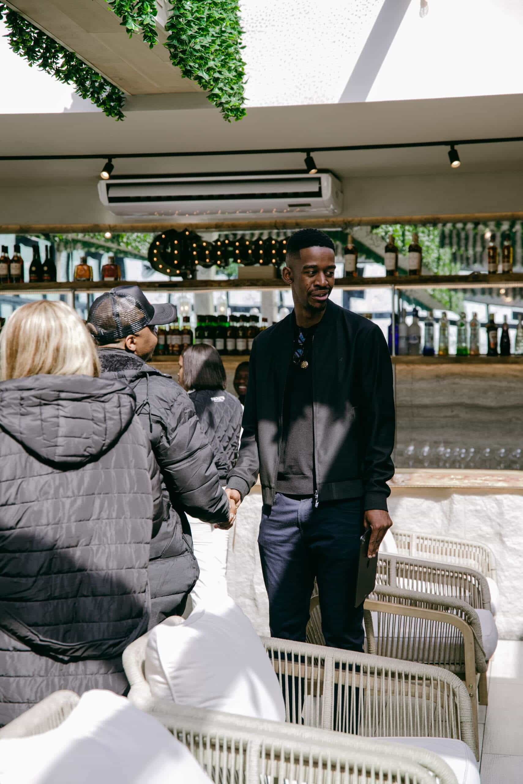 A man in dark clothing stands and smiles near a bar, holding a phone. He is interacting with two people in black jackets, one of whom wears a baseball cap. Indoor area has cushioned chairs, a shelf of bottles, and greenery hanging from the ceiling.