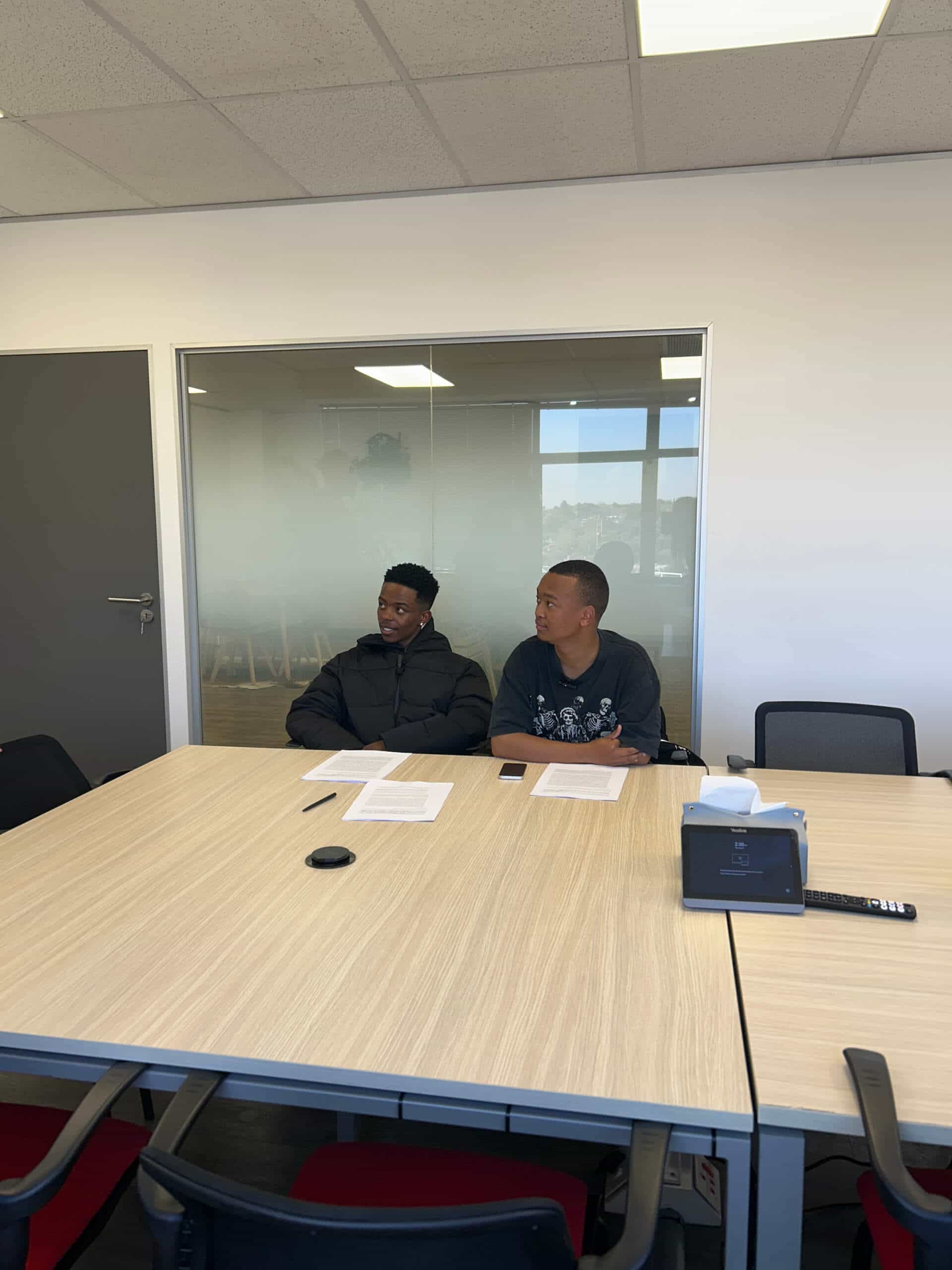 Two men sit at a light wood conference table with papers in front of them, facing slightly to the left. The modern meeting room, part of Downtown Music Publishing Africa&rsquo;s office, features white walls, black chairs, and a visible overhead light.