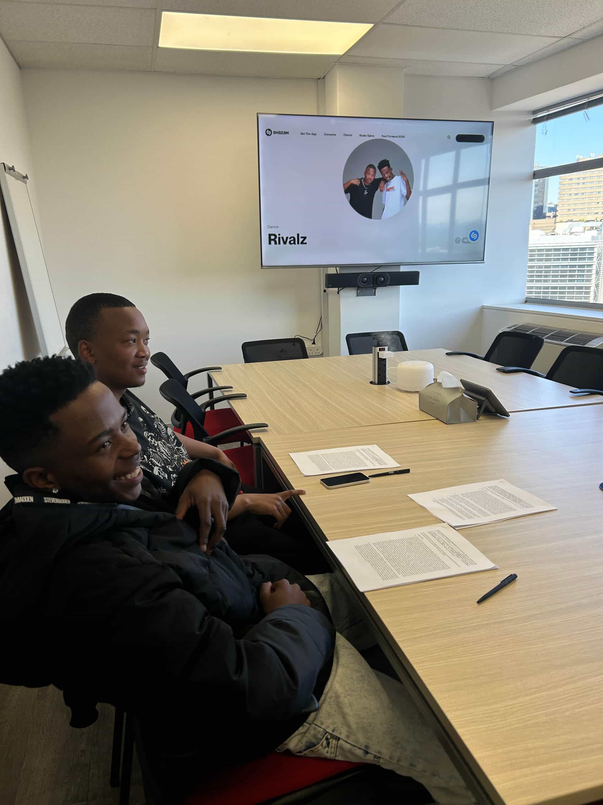 Two smiling people sit at a conference table with papers and pens in front of them. A large screen displays the &ldquo;Rivalz&rdquo; website, featuring three people. The room overlooks the city, with empty chairs and Downtown Music Publishing materials nearby.
