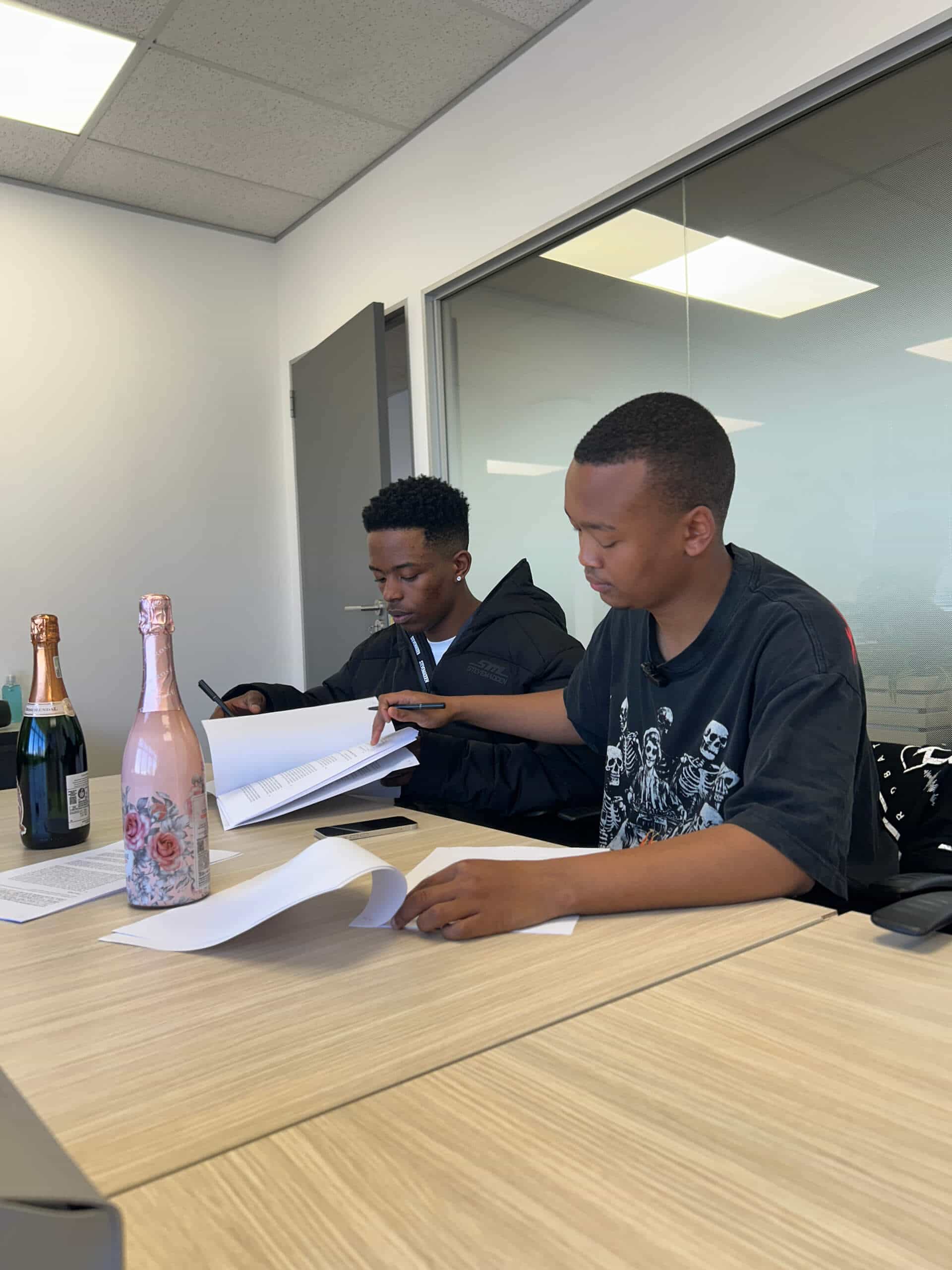 Two young men sit at a wooden table in a modern office, reviewing and signing documents for Downtown Music Publishing Africa. One wears earphones and a jacket, the other a black graphic t-shirt. Bottles of sparkling wine rest on the table beneath fluorescent lights.