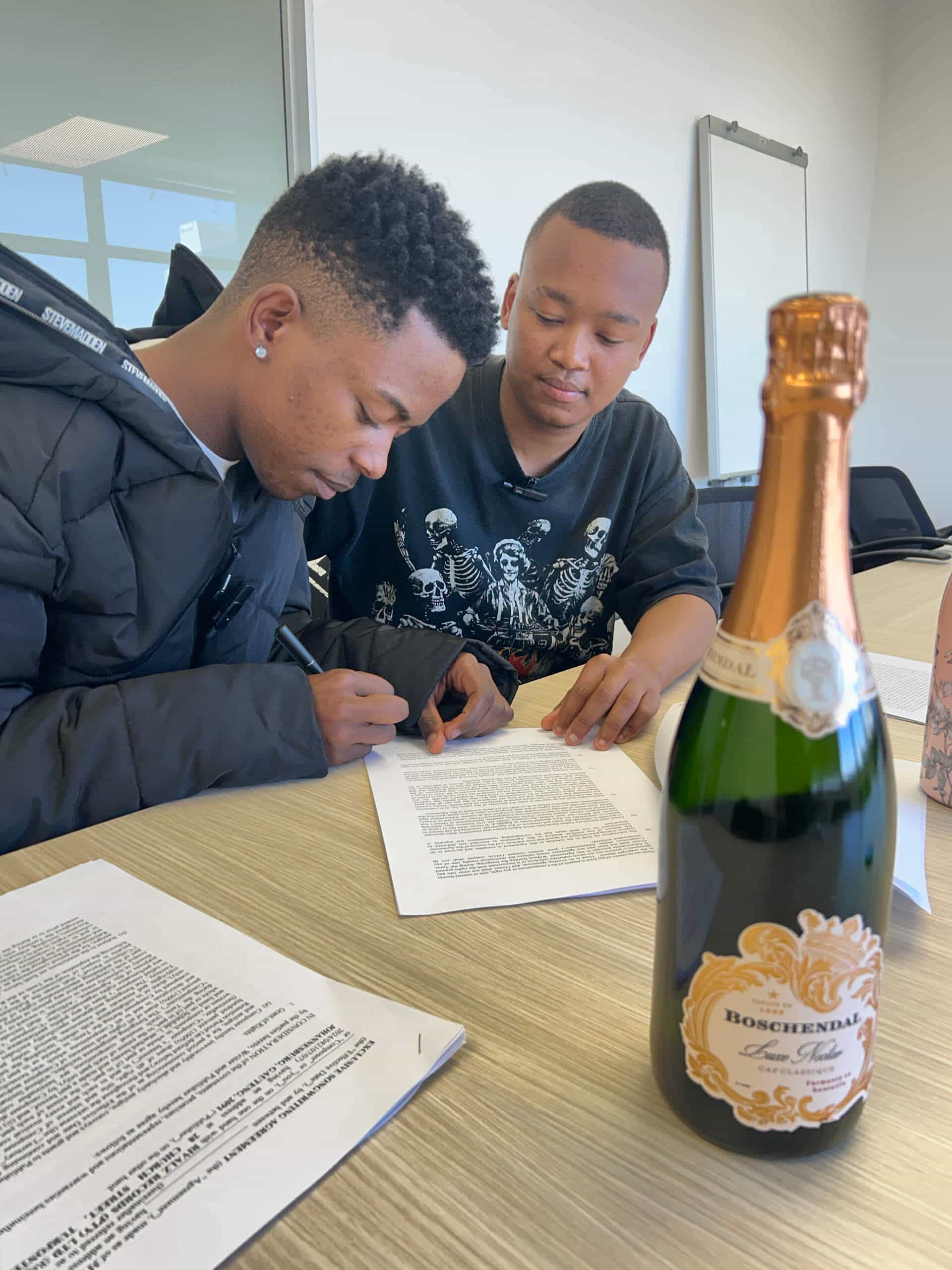 Two young men sit at a table in a bright, modern office, focussing on signing what appears to be a Downtown Music Publishing Africa document. Papers cover the wooden table, with a bottle of Boschendal sparkling wine visible in the foreground.