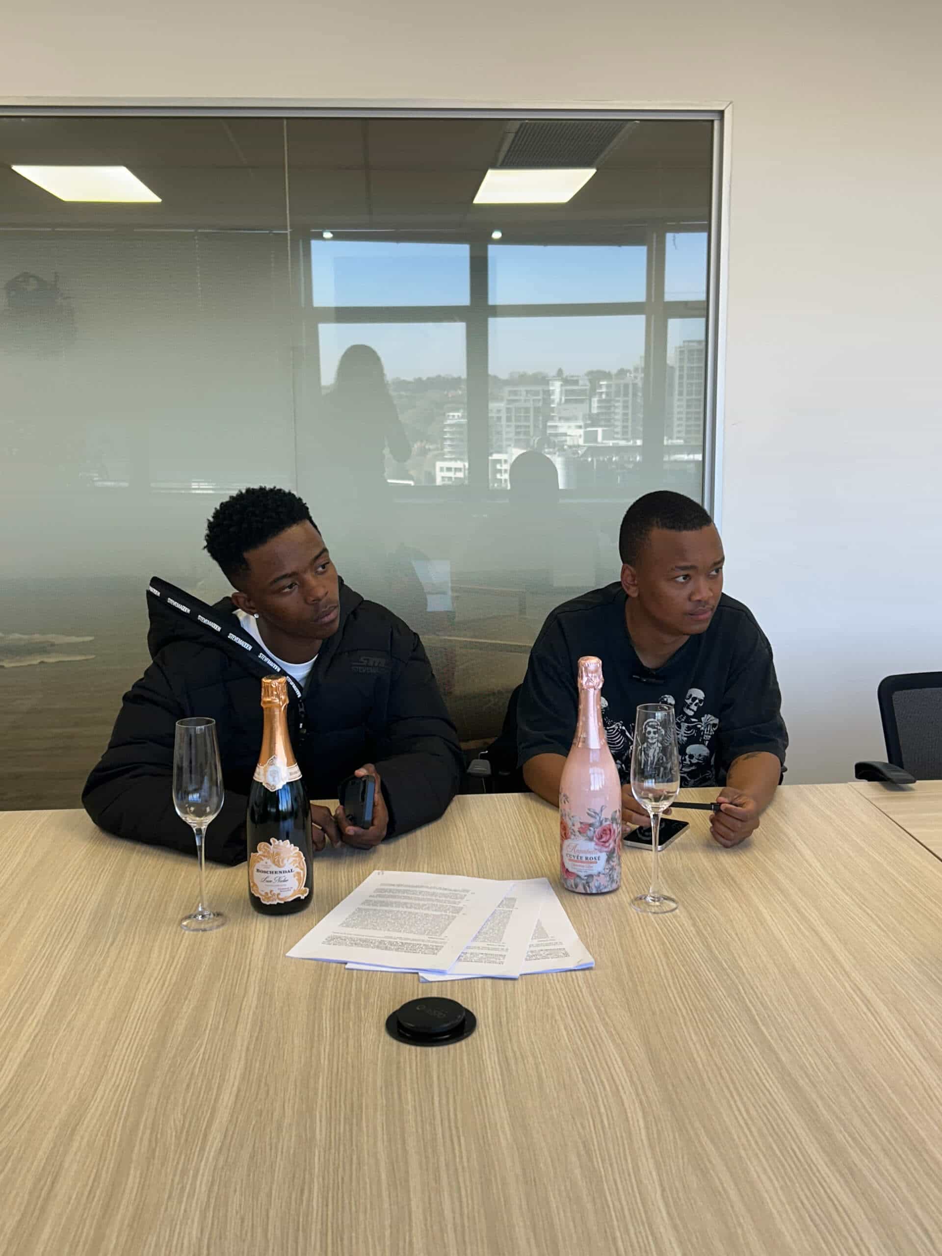 Two men sit at a conference table with champagne bottles and glasses in front of them, papers spread out as they discuss a deal for Downtown Music Publishing Africa. The glass wall behind them reveals an urban cityscape, capturing the Rivalz spirit.