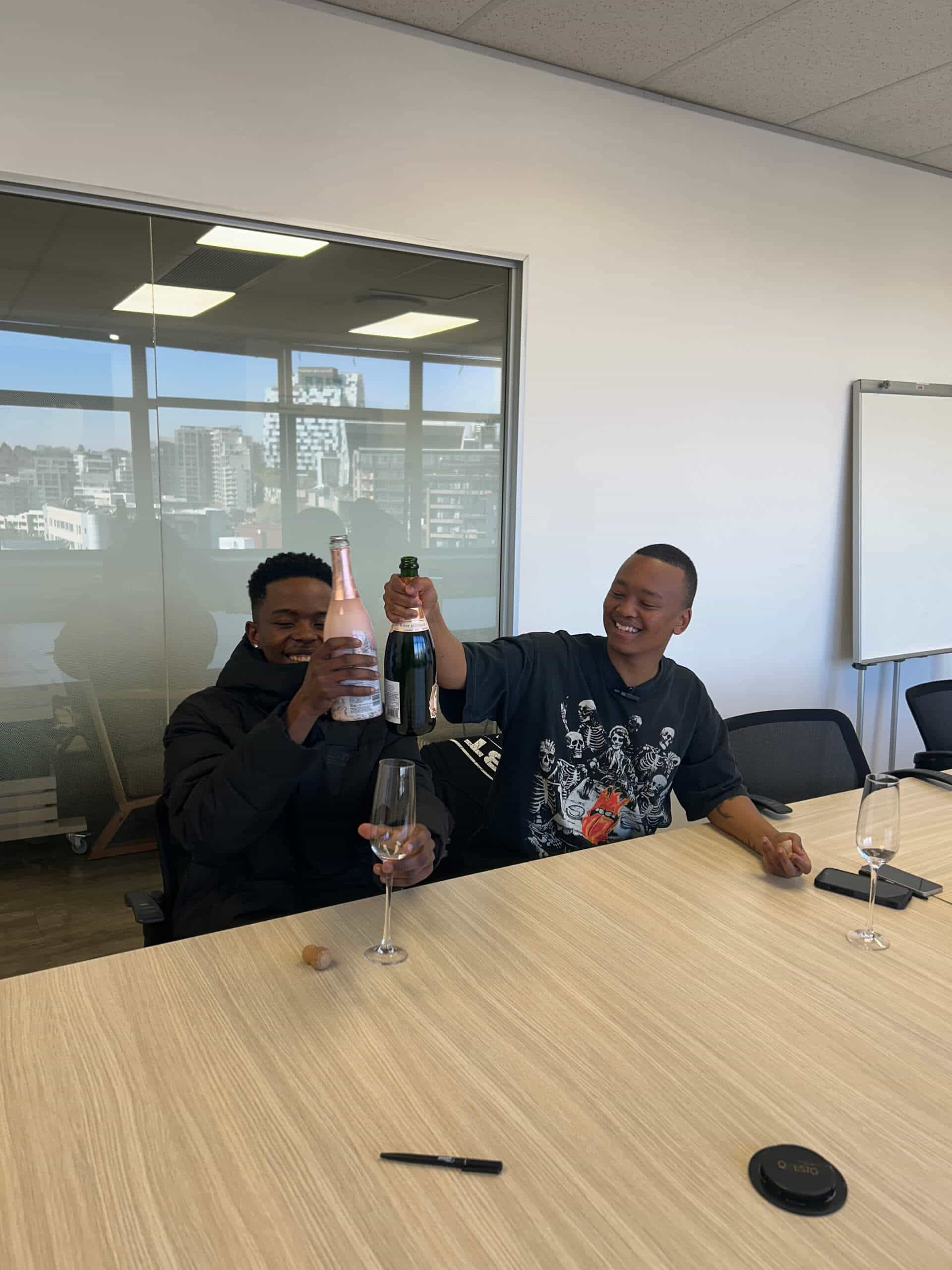 Two people sit at a conference table, laughing and clinking Rivalz bottles of sparkling wine. One holds a pink bottle, the other a green one. Empty glasses and a cork rest on the table, with Downtown Music Publishing Africa&rsquo;s cityscape visible through large windows.