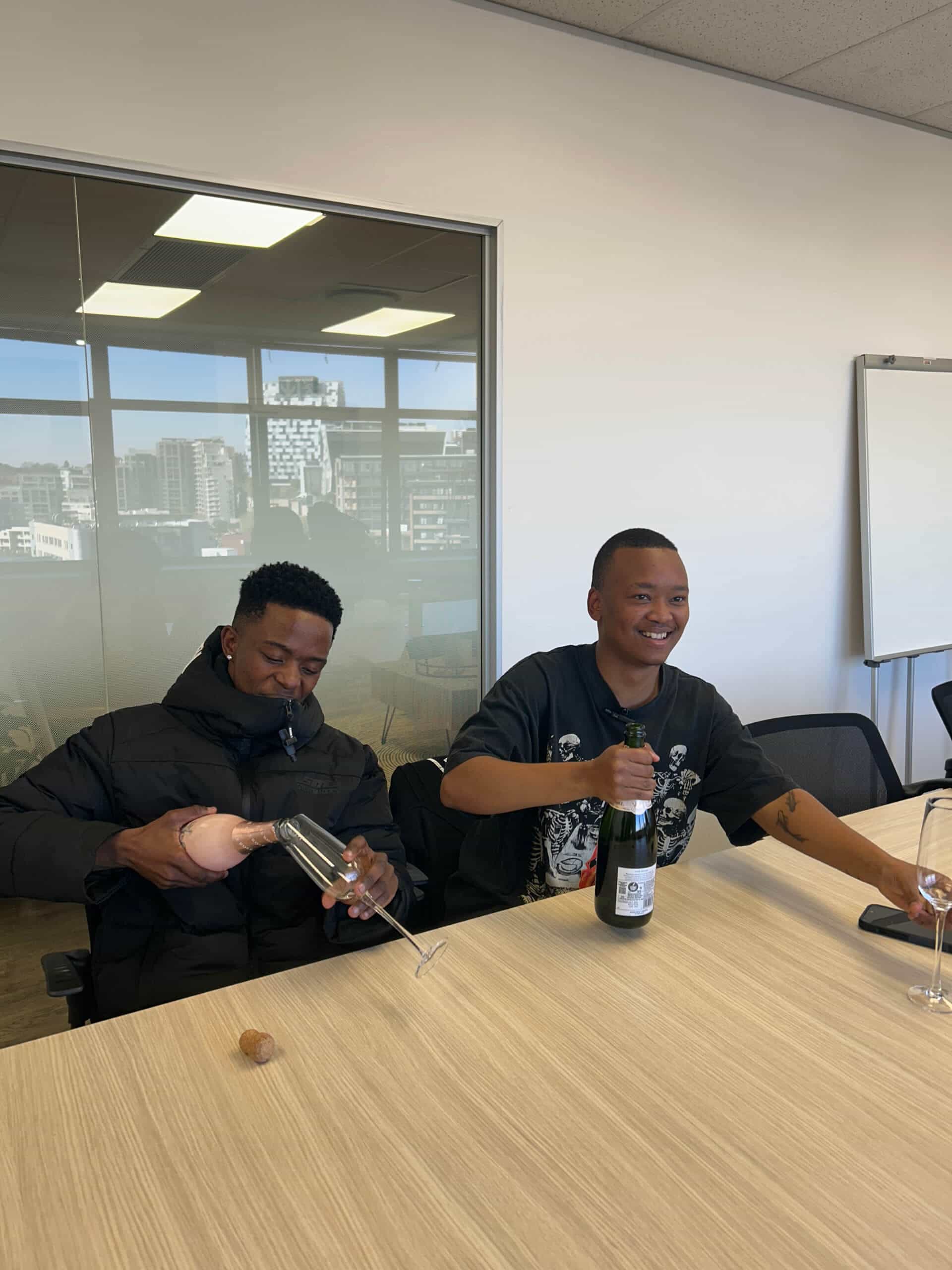 Two people sit at a conference table at Downtown Music Publishing Africa. One, in a black jacket, pours ros&eacute; wine into a glass, while the other, wearing a Rivalz graphic T-shirt, opens sparkling wine. Office windows and city buildings are visible behind them.