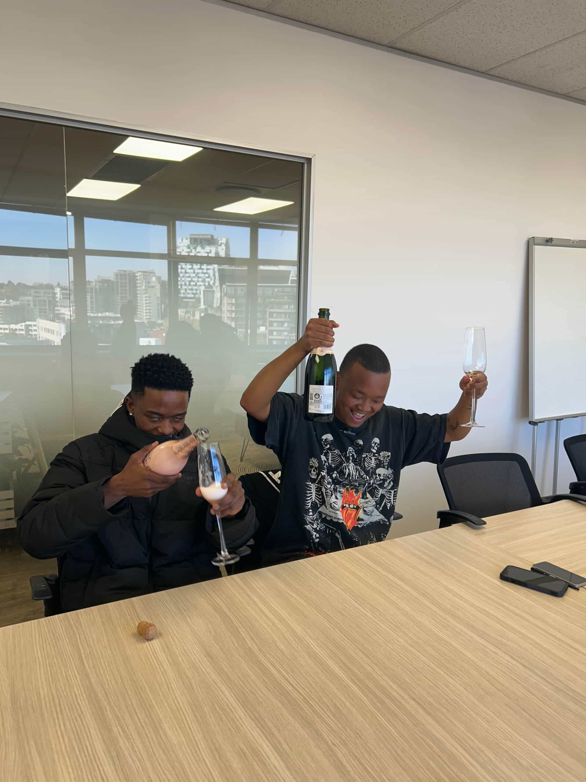 Two men sit at an office table. The man on the left, pouring sparkling wine into a flute, wears a black jacket. The man on the right, smiling widely, celebrates with Downtown Music Publishing Africa as city buildings rise behind them.