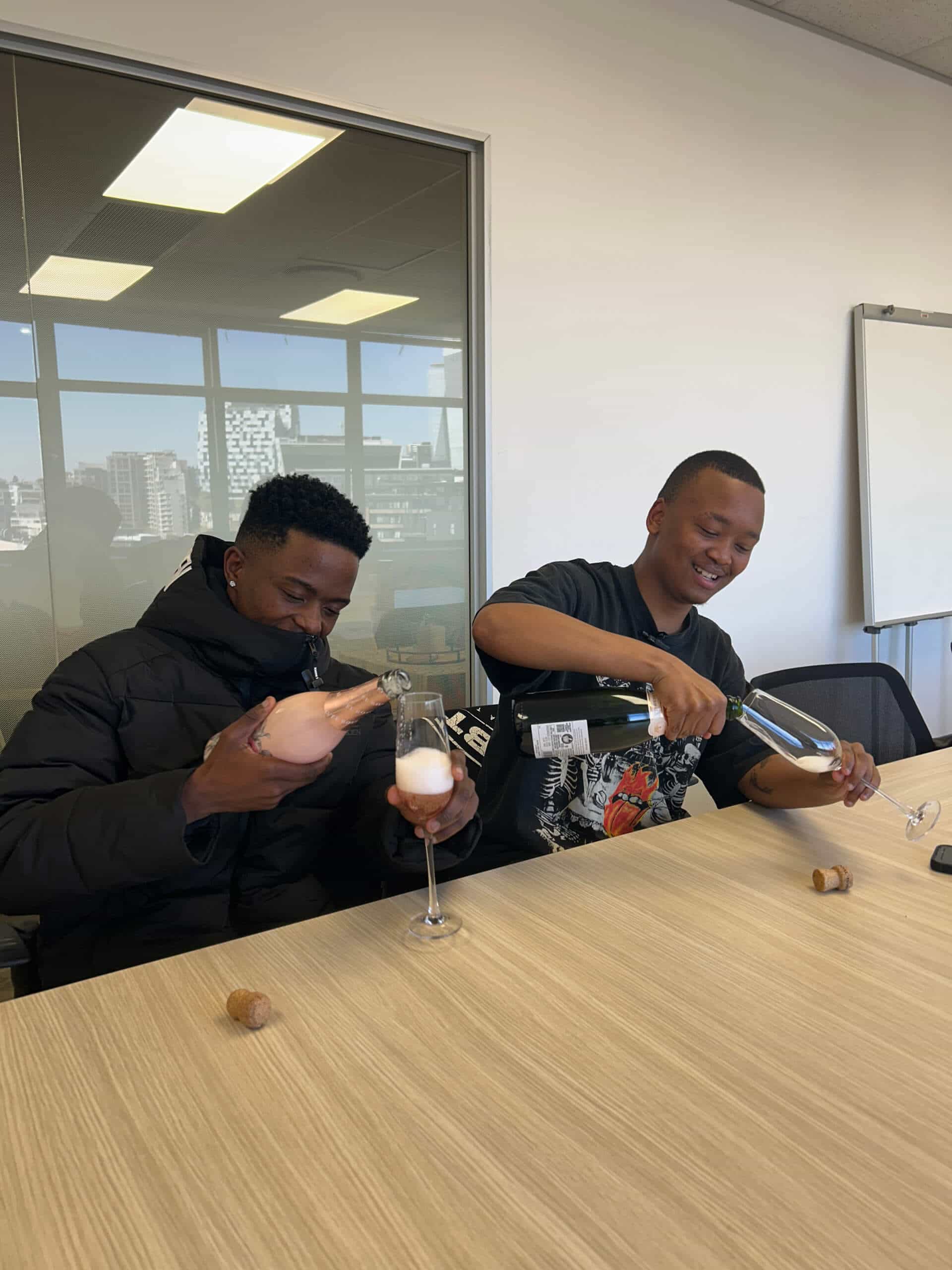 Two young men sit at a wooden table, each pouring sparkling wine into tall glasses. Laughter fills the air as Rivalz plays softly; champagne corks lie on the table, and a cityscape hints at Downtown Music Publishing Africa beyond the window.
