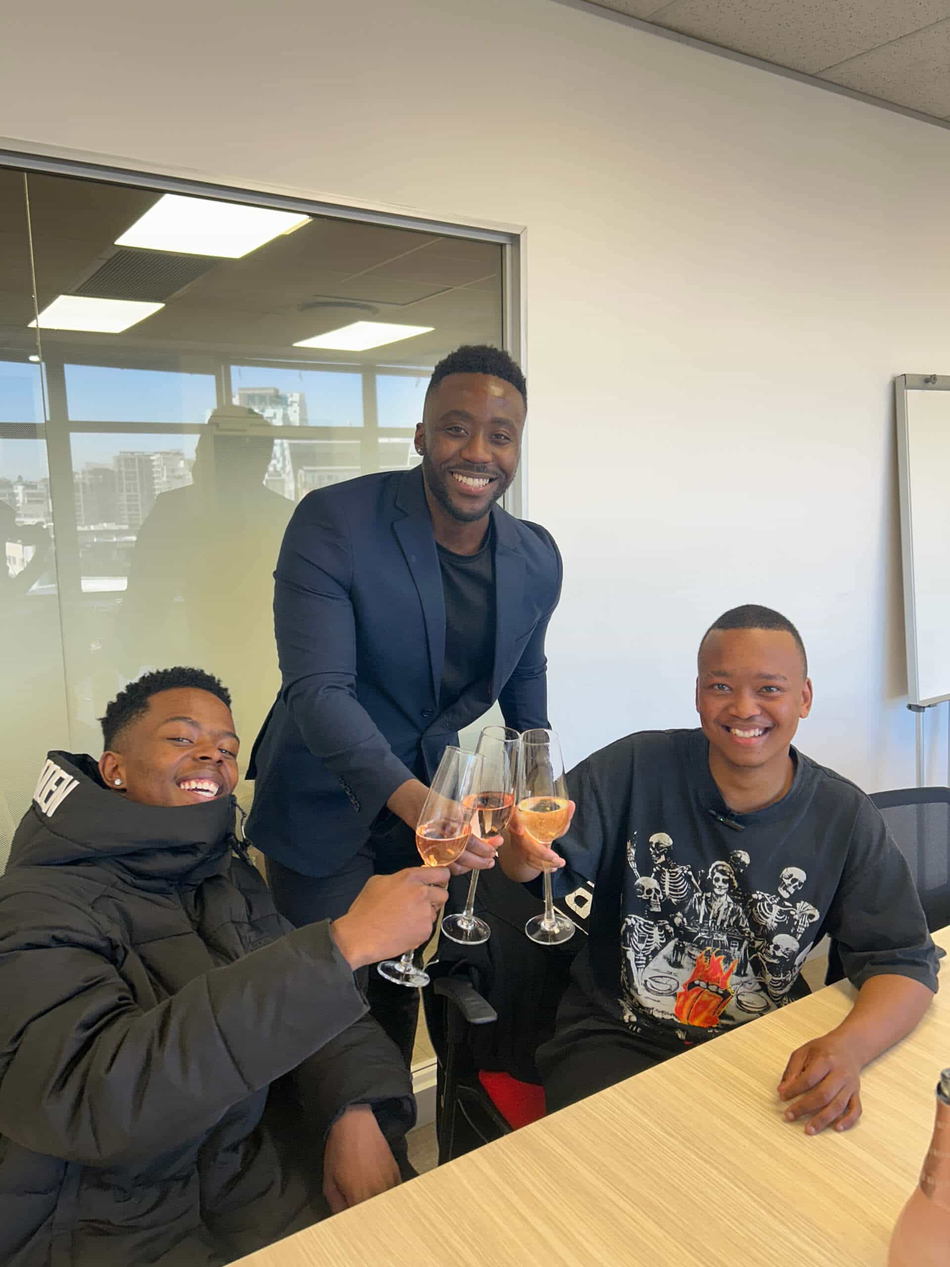Three men smile and toast with glasses of ros&eacute; wine in a modern office. One wears a Rivalz graphic T-shirt, while another sports a navy blazer. Gathered at a wooden table, they celebrate against the backdrop of Downtown Music Publishing Africa.