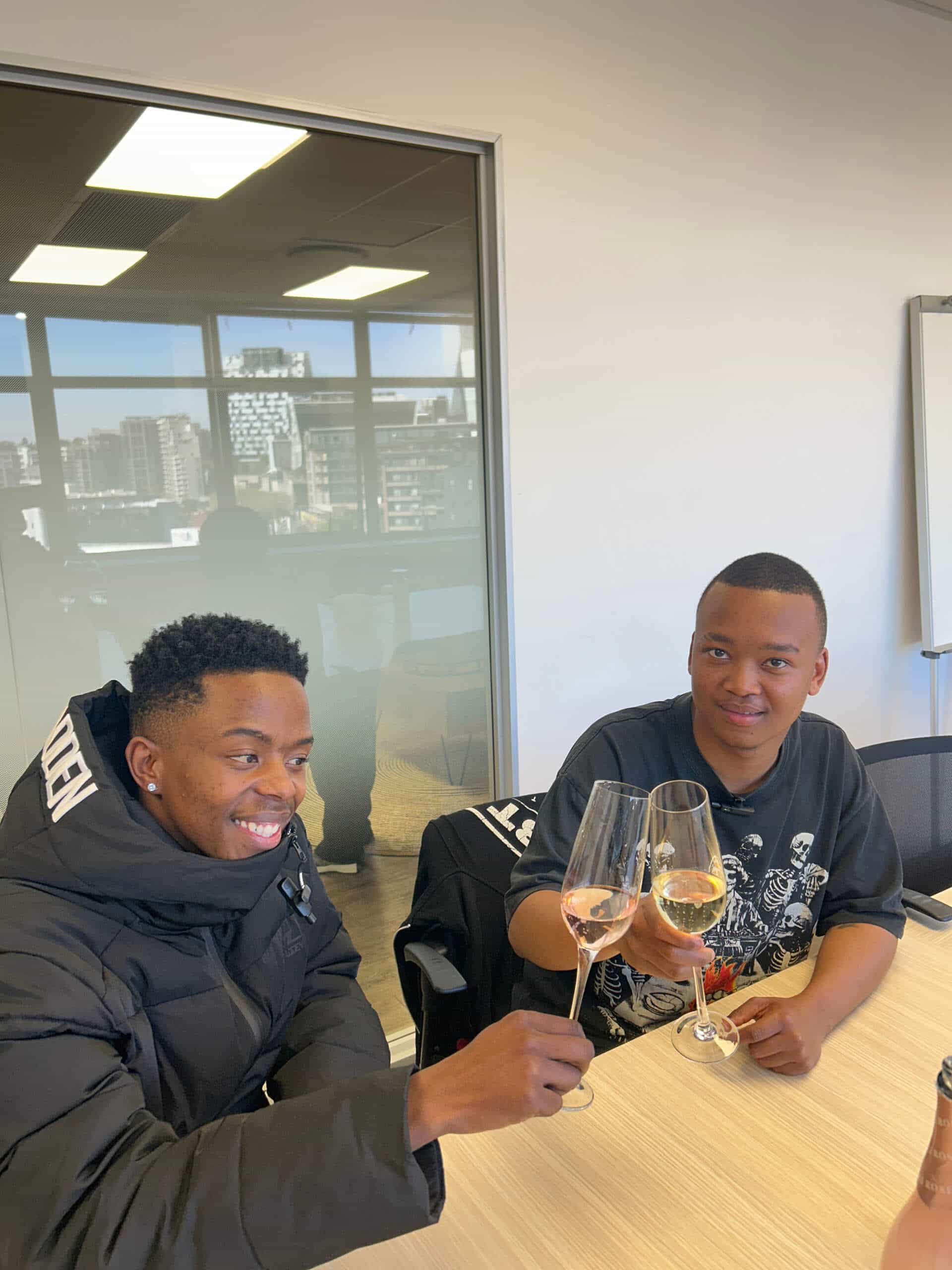 Two young men sit at a table in an office, smiling as they toast with glasses of ros&eacute; wine. One wears a black puffer jacket, the other a black graphic T-shirt. Behind them, Downtown Music Publishing Africa&rsquo;s cityscape and glass-walled meeting room gleam.