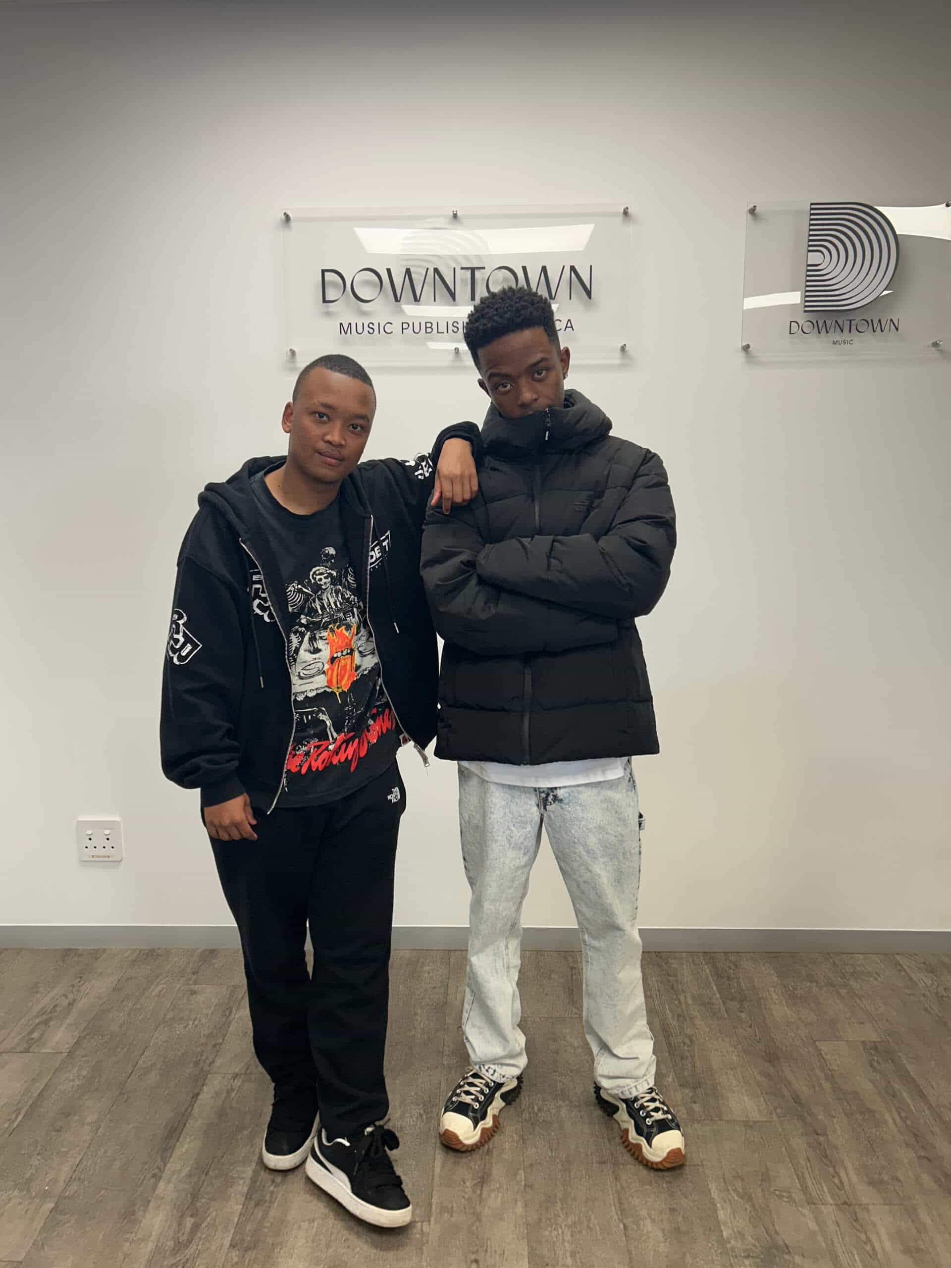 Two young men stand indoors in front of &ldquo;DOWNTOWN MUSIC PUBLISHING&rdquo; signs. One wears a black hoodie, the other a puffer jacket. Representing Rivalz, they're pictured at Downtown Music Publishing Africa, friendship and music uniting them.