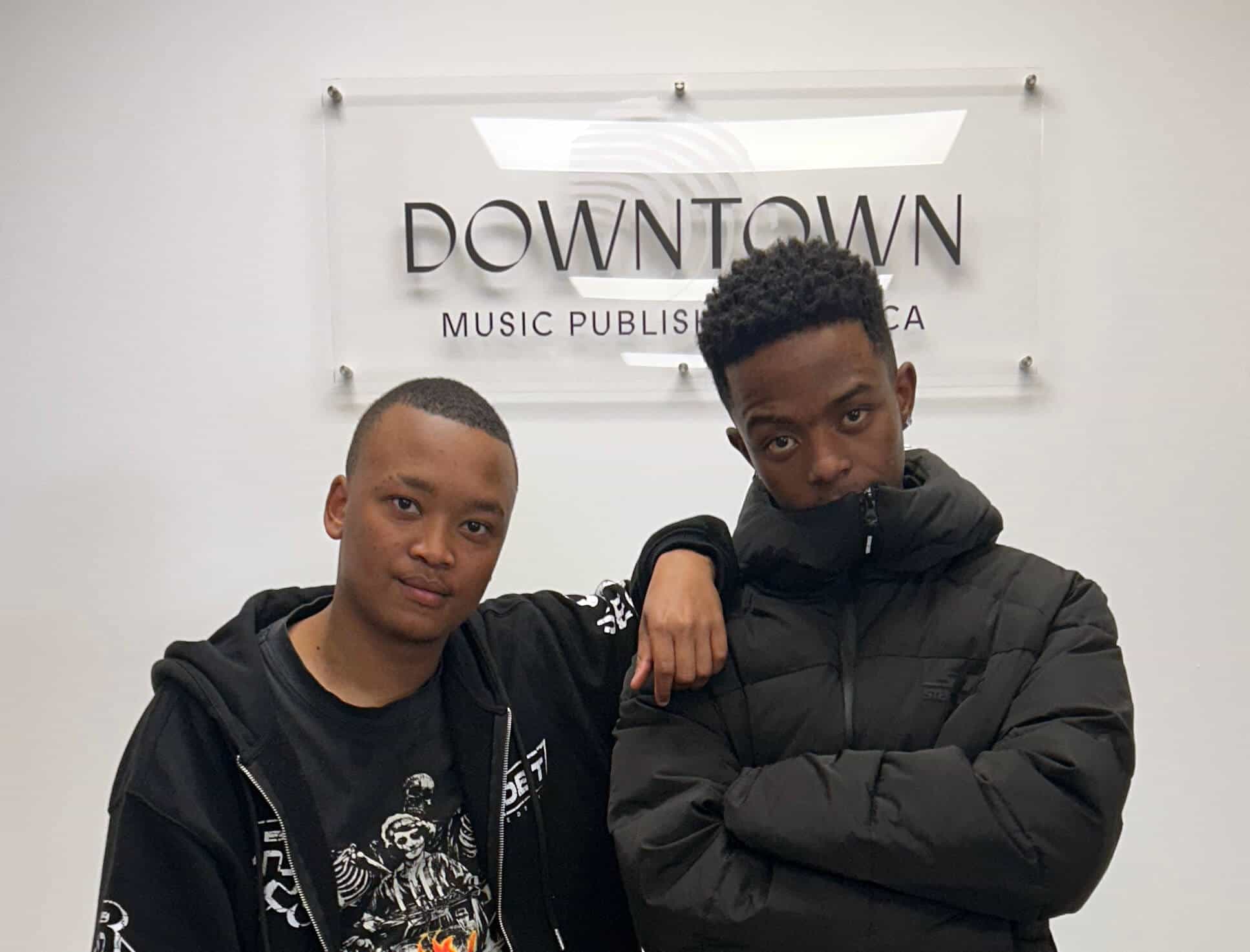 Two young men, including Rivalz, stand by a wall with a &ldquo;Downtown Music Publishing Africa&rdquo; sign&mdash;one in a black graphic hoodie with his arm round the other in a puffer jacket. Both look at the camera with relaxed, serious expressions.