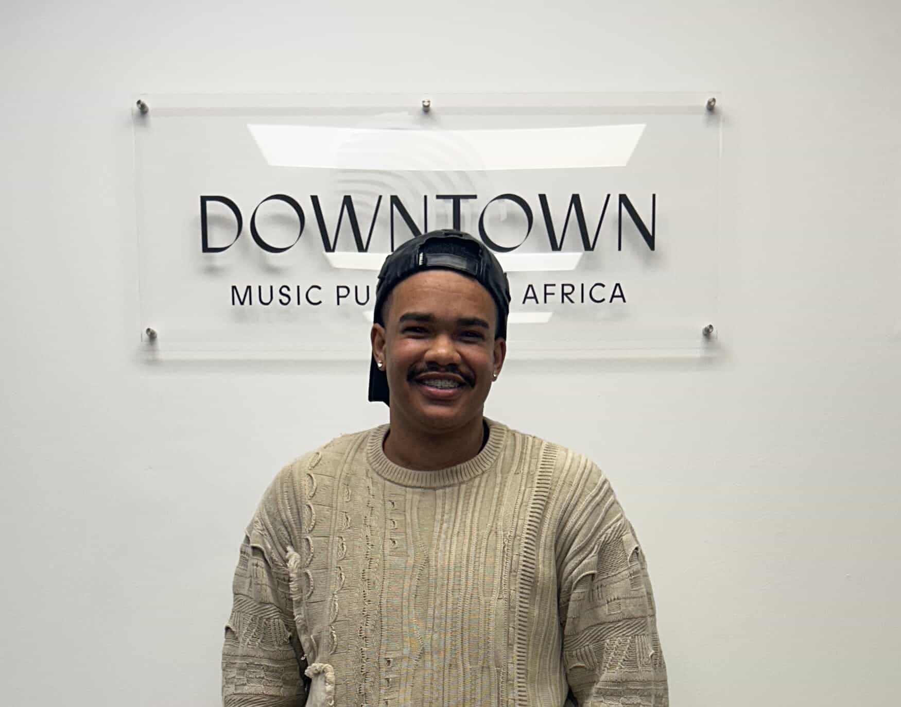 A person with a moustache and short hair, wearing a textured beige jumper and a backwards black cap, smiles whilst standing in front of a wall sign that reads "Downtown Music Publishing Africa," celebrating their latest publishing deal.